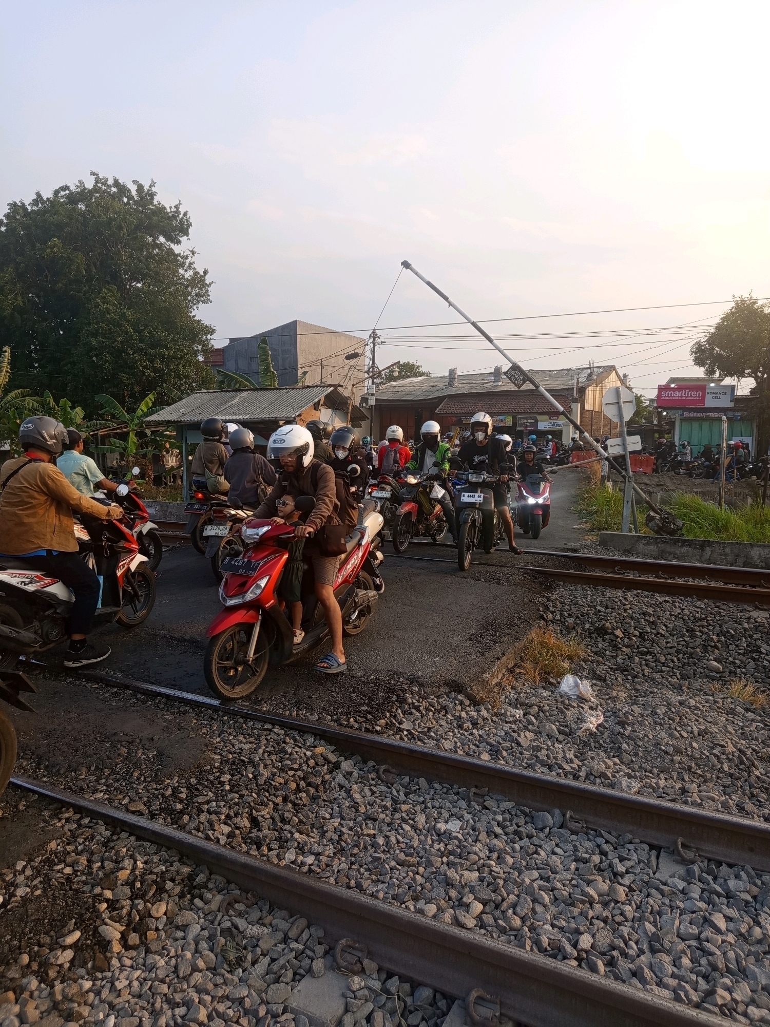 The image captures a busy street scene at a railway crossing in Semarang, Indonesia. Several people on motorcycles are waiting at the crossing, suggesting a pause in traffic. The crossing gate is partially lowered, indicating an approaching train or that the crossing is currently active. The environment appears to be a typical urban setting with buildings and trees visible in the background. The lighting suggests it is either early morning or late afternoon. The railway tracks are prominent in the foreground, with gravel ballast typical of such infrastructure. The people are dressed casually, with many wearing helmets, which is common practice for motorcycle riders in Indonesia. Some motorcycles have passengers, including a child riding with an adult. The overall impression is one of everyday life in a bustling Indonesian city.