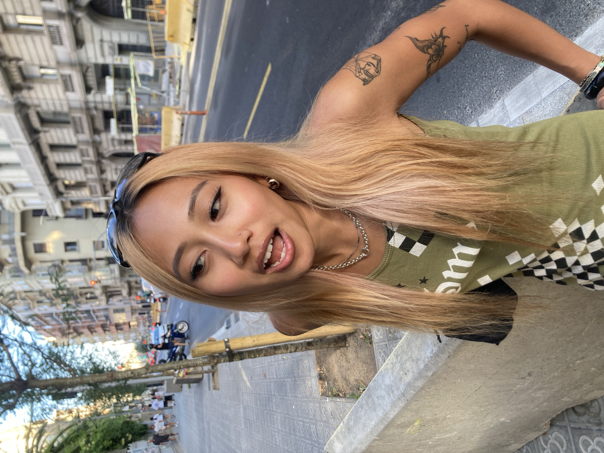 The image is a close-up, rotated selfie of a young person with blonde hair, taken outdoors in Barcelona, Spain. The subject is wearing a green tank top with a checkered pattern and has several tattoos on their right arm, including one that appears to be a spider web with a face in the center and another of a butterfly-like design. They are also wearing a silver necklace and hoop earrings. The person is looking directly at the camera with their mouth slightly open, appearing to be speaking or making a playful expression.
In the background, a city street is visible with buildings, traffic, and trees. The architecture of the buildings suggests an urban setting, and the presence of traffic lights and road markings confirms it's a street scene. The lighting suggests it's daytime, possibly late afternoon due to the soft light. There are also some construction barriers and scaffolding in the background, indicating ongoing work. Further in the background, other people can be seen walking along a sidewalk and sitting at outdoor tables, hinting at a lively urban environment. The overall scene evokes a casual, everyday moment captured during a day out in the city.