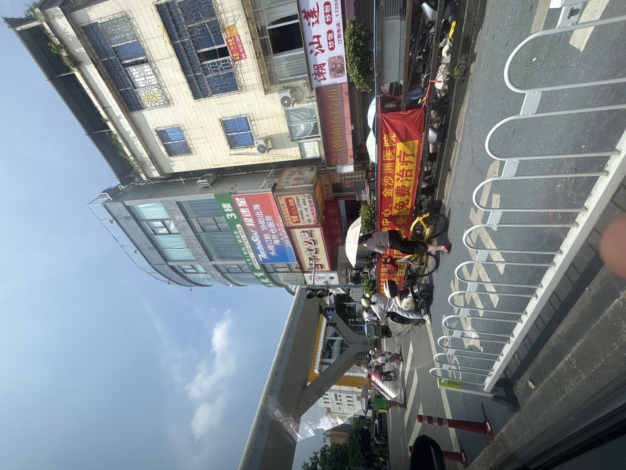 In Baiyun District, China, a street scene unfolds under a bright sky with scattered clouds. A multi-story building with a curved facade dominates the right side of the image, featuring a rooftop garden and numerous windows with metal grates. Adjacent to it, a lighter-colored building displays similar barred windows and air conditioning units.
In the foreground, a white metal barrier runs along the side of a street. Traffic is present, with several motorcycles and a bicycle visible. One person rides a motorcycle, while another sits on the back of a scooter, both wearing helmets. A person on a bicycle, wearing a striped shirt and dark shorts, holds a light-colored umbrella.
A prominent red banner stretches across the scene, displaying yellow Chinese characters that translate to "Center, Jinsha Hospital, Free Treatment." The banner is supported by a cart or a vehicle. Orange and white traffic cones are placed on the street. An elevated walkway or bridge structure is visible in the background, spanning across the street. Businesses are advertised on the buildings, with signs in Chinese. The overall impression is a bustling urban environment with a focus on a medical promotion.
