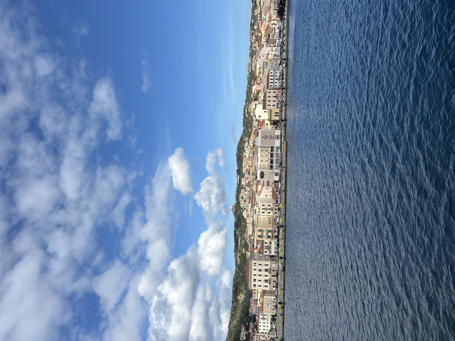 The image displays a panoramic view of the coastal town of La Maddalena, Italy, under a partly cloudy blue sky. The scene is bisected horizontally by the horizon line of the water and the town.
The upper portion of the image features a bright blue sky, predominantly clear on the right side, transitioning to scattered, fluffy white cumulus clouds on the left.
The lower portion of the image is dominated by deep blue-green water, exhibiting ripples and small waves across its surface, suggesting a gentle current or breeze. No watercraft are visible.
Above the water, the town of La Maddalena stretches horizontally, fronted by a paved waterfront promenade or quay. Numerous parked vehicles, mostly dark-colored sedans, are visible along this waterfront road, accompanied by yellow bollards or barriers in sections. Behind the quay, a dense collection of multi-story buildings forms the immediate urban area. These structures vary in color, including shades of cream, pastel yellow, pink, and light green, with some darker facades. Architectural styles appear typical of Mediterranean port towns. A prominent light-colored building with multiple floors and arched windows is situated towards the left-center of the waterfront.
Behind the waterfront buildings, the town ascends the slopes of a green, rocky hill. More residential and commercial structures are visible further up the hillside, integrated into the natural terrain which is covered by sparse vegetation and exposed rock. Near the highest visible point of the hill, towards the left, a distinct white structure featuring a spire or dome is discernible, identified as a church or chapel (likely the Santuario della Madonna di Lardari).
No discernible human figures or specific actions are visible within the frame, indicating a static, distant view of the town and its environment.