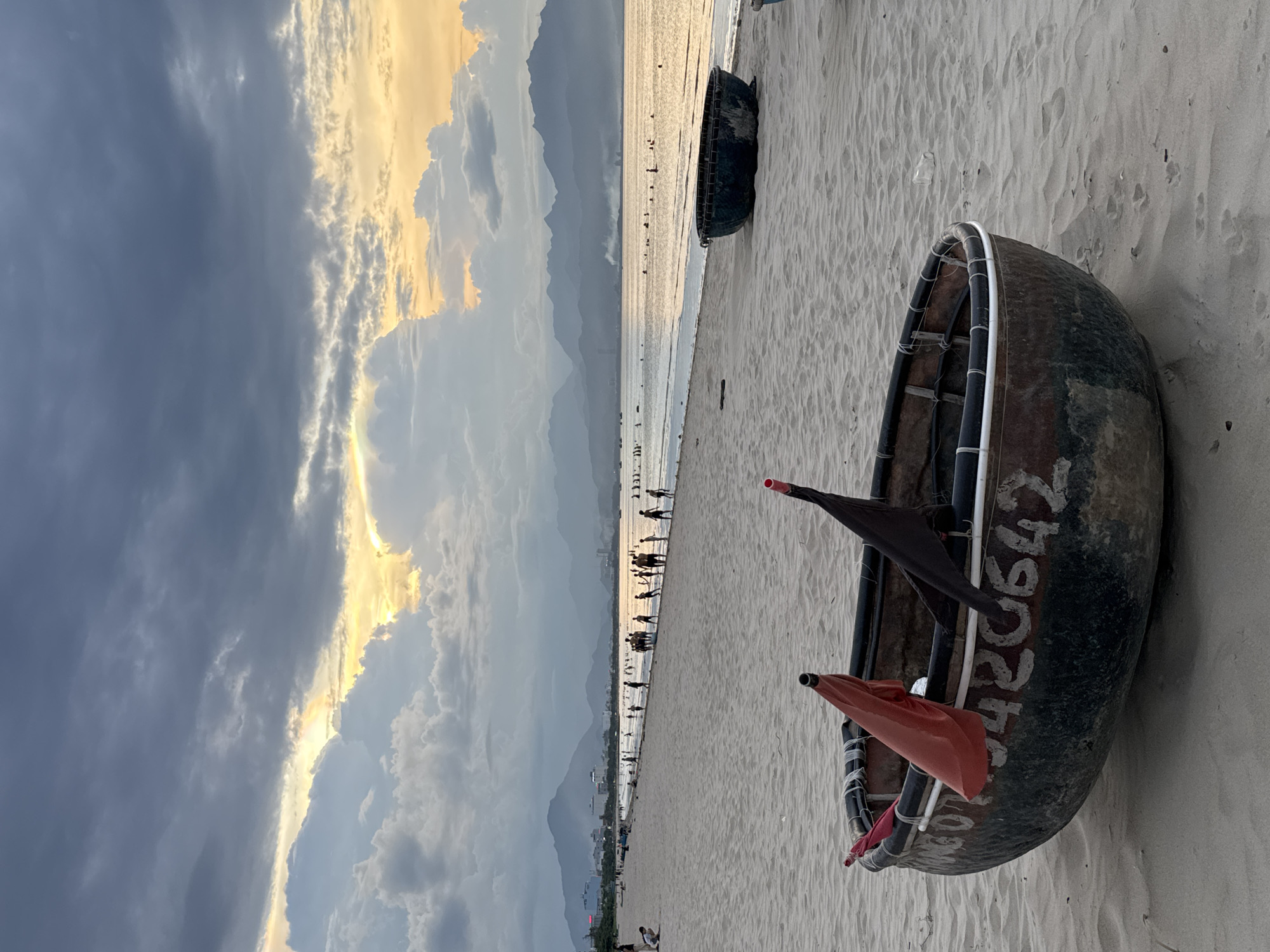 This is a vertical, eye-level medium shot of a Vietnamese basket boat resting on a sandy beach at sunset. The boat, a round, dark-colored vessel, is positioned in the foreground on the right side of the frame. It has white numbers "420642" painted on its side. Two small flags, one orange and one black, are tied to its rim. The beach is made of light-colored sand, with footprints visible.
In the background, the calm ocean stretches out, reflecting the warm hues of the setting sun. A thin line of people can be seen walking along the shoreline, suggesting a public beach. In the far distance, a range of mountains creates a silhouetted backdrop against the sky. The sky itself is dramatic, with dark, moody clouds on the left, interspersed with streaks of golden light from the setting sun on the right, indicating a transition from day to evening. The overall atmosphere is serene and tranquil, with a sense of natural beauty. The location is identified as Đà Nẵng, Vietnam.