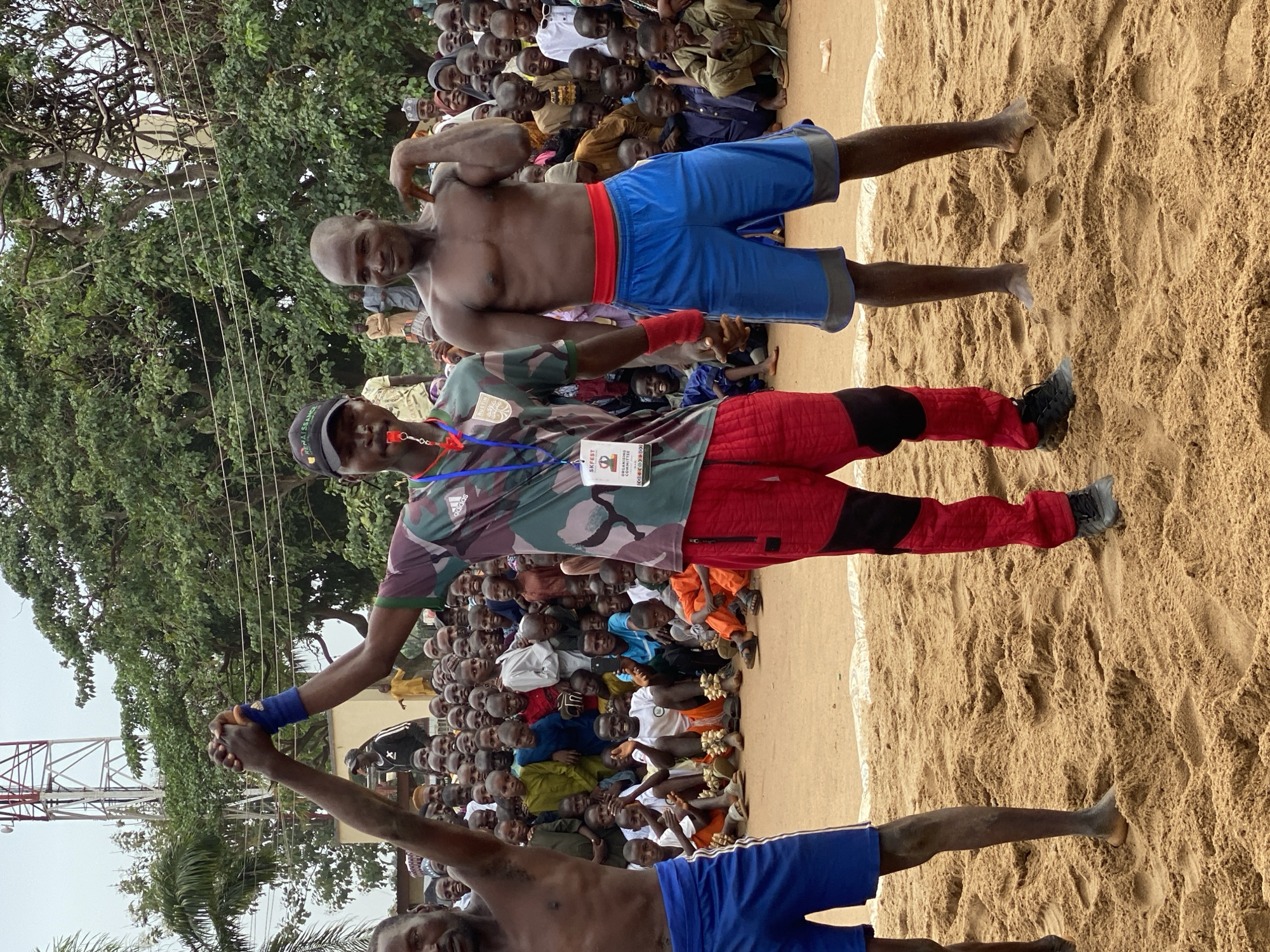 The image depicts a scene from what appears to be a traditional wrestling event. Two bare-chested men, each wearing blue shorts with red waistbands, stand on a sandy surface. A third man, wearing a camouflage shirt, red pants, and a baseball cap, stands between them, possibly acting as a referee or official. He is wearing a lanyard.
A large crowd of spectators surrounds the wrestling area. The location appears to be outdoors, with trees visible in the background. A cell tower is also visible in the background. It has been suggested that this image was taken in Kafanchan A, Nigeria.