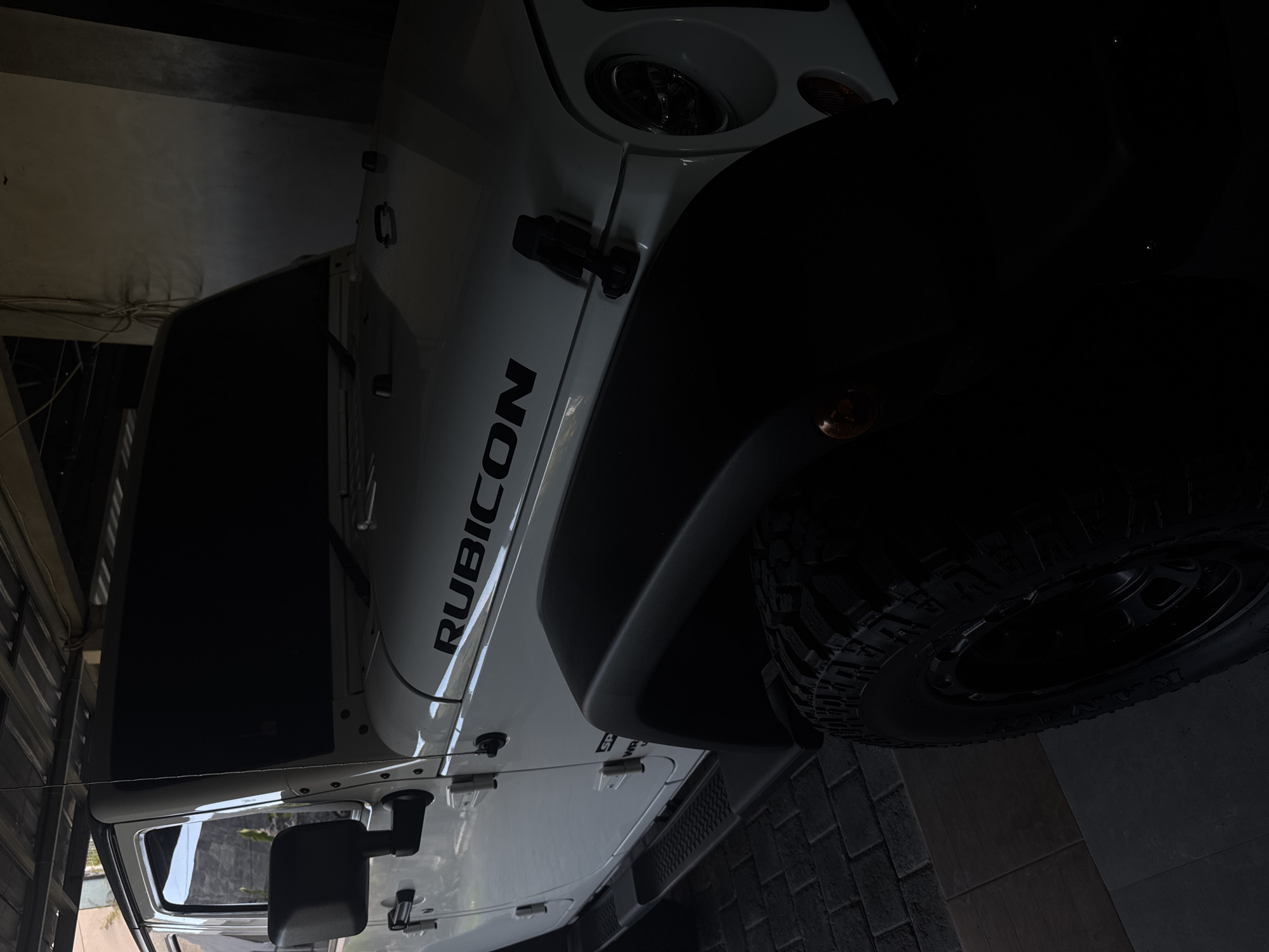The image depicts a close-up view of a white Jeep Rubicon, with the word "RUBICON" prominently displayed on the side. The vehicle is parked on a brick surface. The photo was taken indoors, with low lighting, possibly in a garage or a covered parking space. It is daytime. There is no mention or visual evidence to suggest the location is in North Kuta, Indonesia. The image focuses on the details of the Jeep's design. No people, specific actions, or interactions are visible in the image.