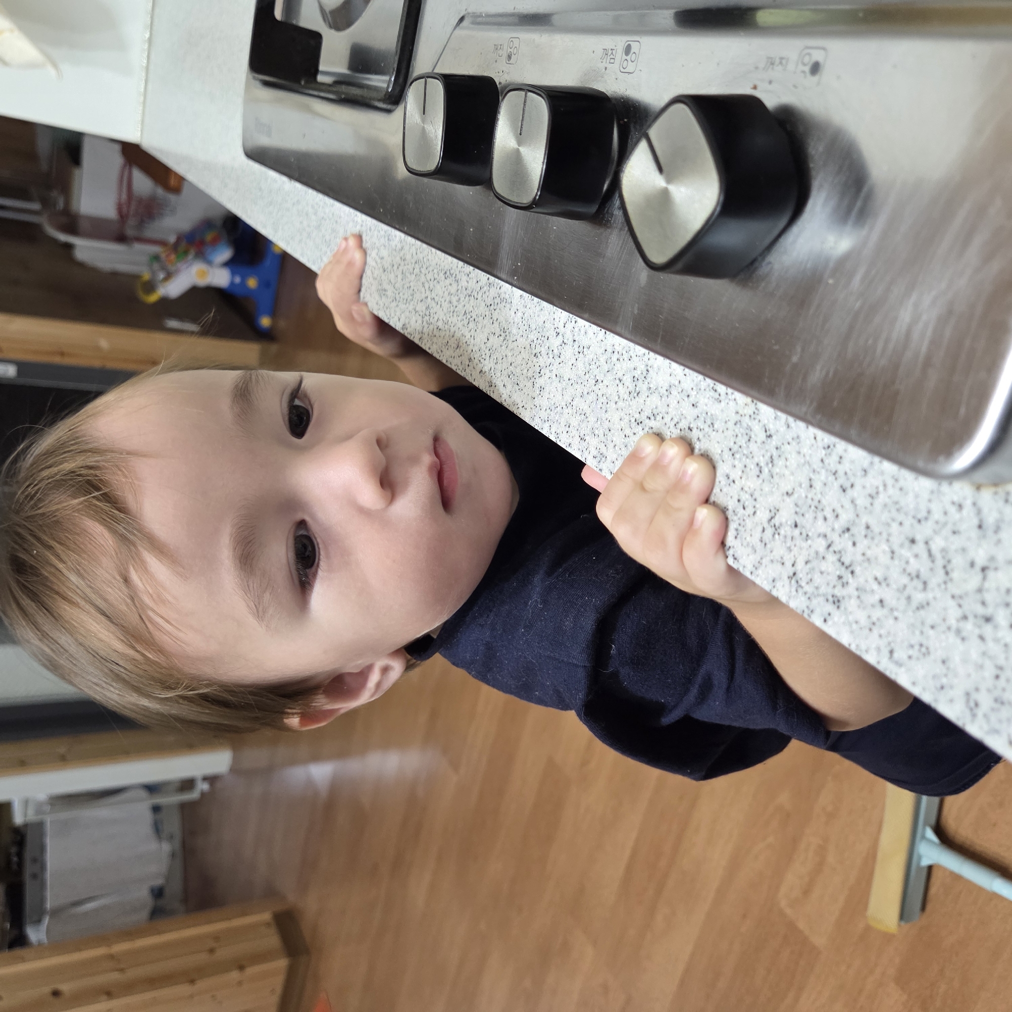 A toddler stands on a wooden floor, leaning on a speckled countertop with their hands. They are wearing a dark blue long-sleeved shirt and looking down at the countertop. In the background, a child's toy is visible, along with wooden paneling on a wall and what appears to be kitchen cabinetry. The toddler's position suggests they are in a home environment, possibly a kitchen. The specific location of Dunnae-myeon, South Korea, cannot be determined from the visual information provided in the image.