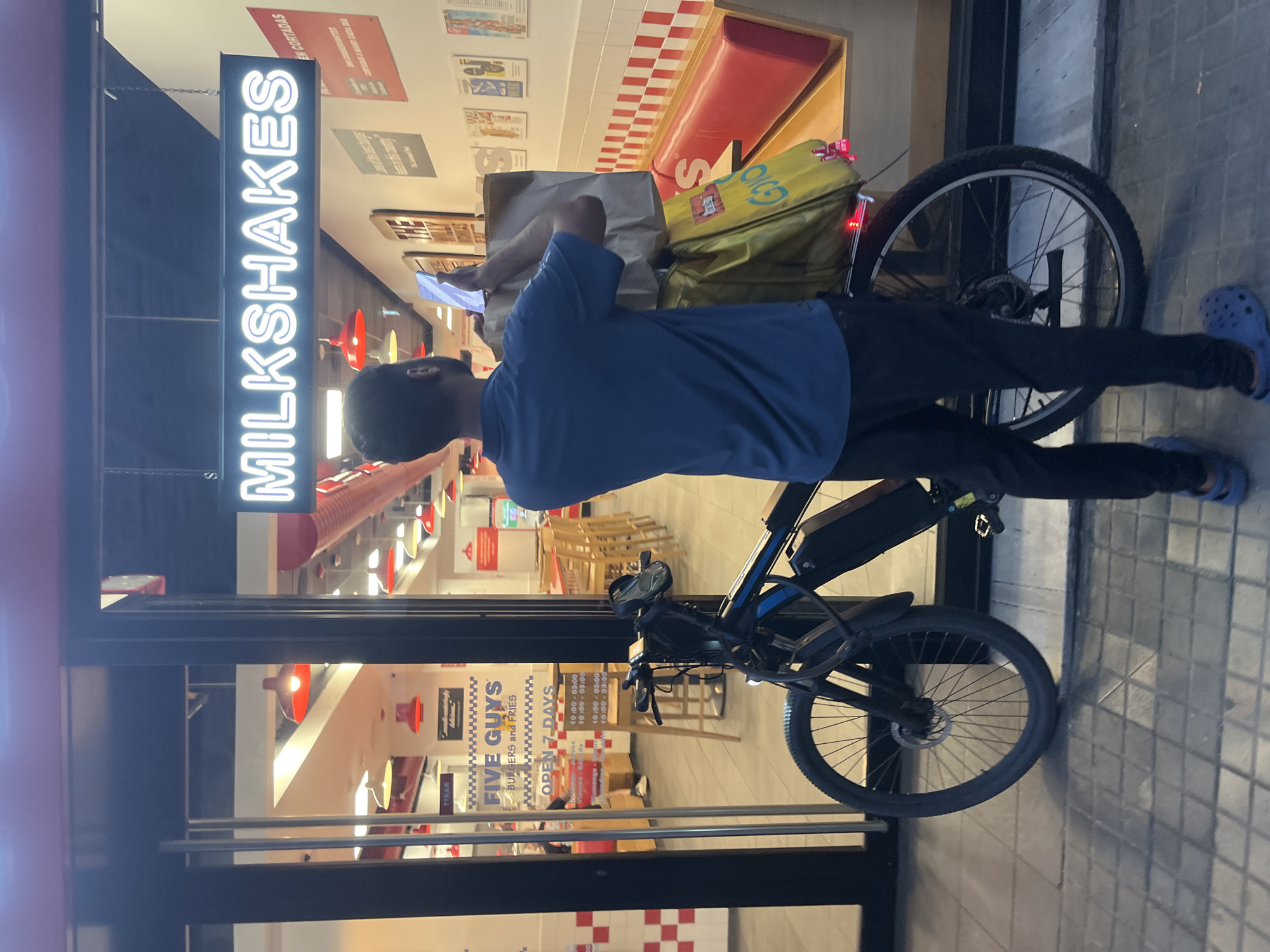 A person is standing next to an electric bicycle in front of a Five Guys restaurant in Barcelona, Spain. The person is wearing a blue polo shirt and black pants, and blue Crocs. They are holding a cell phone and a takeout bag. The electric bicycle has a yellow delivery bag and a brown paper bag attached to the rear rack. The Five Guys restaurant has red and white checkered tiles and red booths. A neon sign above the entrance reads "MILKSHAKES". The street is paved with grey tiles.