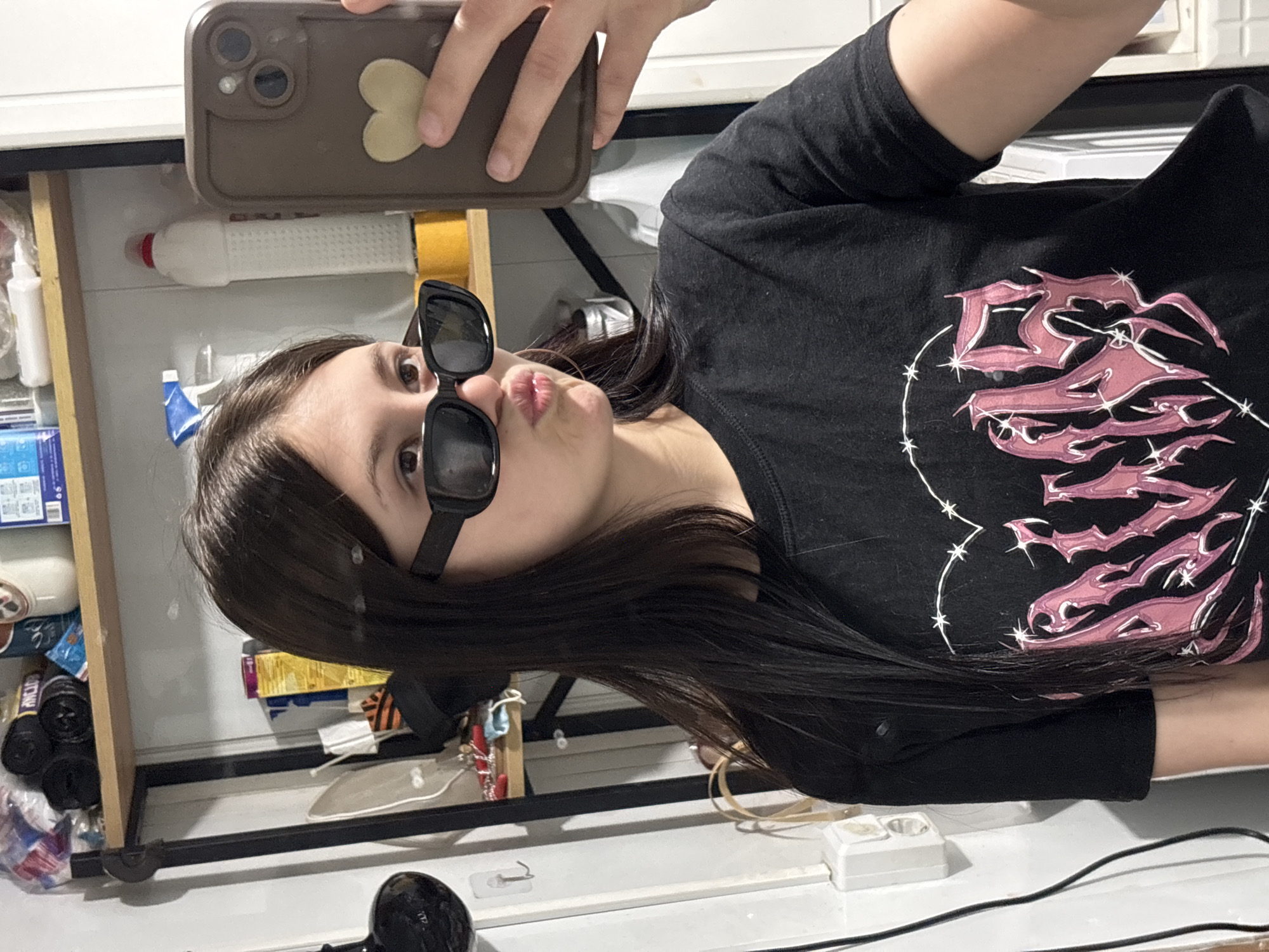 A young woman is taking a selfie. She's wearing black sunglasses and a black long-sleeved shirt with a pink graphic design. She is holding a cell phone with a brown case. The background features a partially opened cabinet with various household items inside. A hairdryer sits on a white counter. The location is impossible to determine from the image.