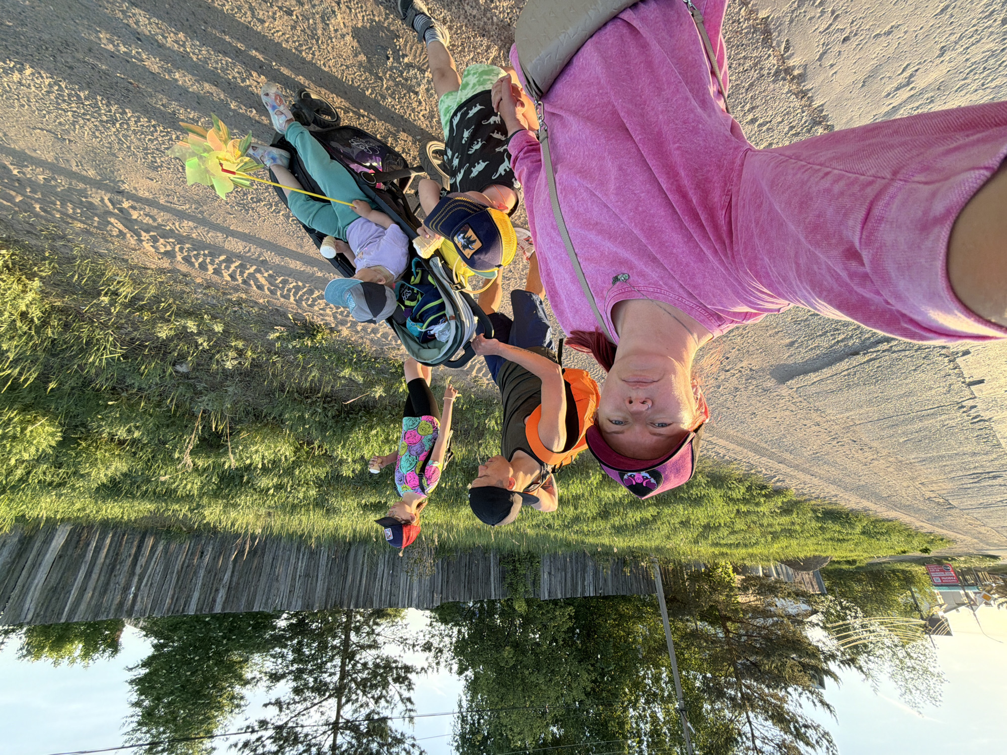 The image captures a family scene on what appears to be the outskirts of Chaikovsky, Russia, judging by the presence of characteristic wooden fences and signage in Cyrillic visible in the background. A woman in a pink top and backwards pink cap is holding the camera, likely taking a selfie. She is joined by a boy with a black and red cap and a girl in a smiley face patterned shirt, who are standing close to her. A man in an orange vest and black cap stands in the middle of the group. In front of them is a stroller, occupied by a toddler holding a pinwheel and a baby bottle. A young boy in dinosaur-patterned shorts is standing by the stroller. The group is positioned on a sandy dirt road, bordering a grassy area with trees. The image orientation is rotated, giving an upside-down perspective.