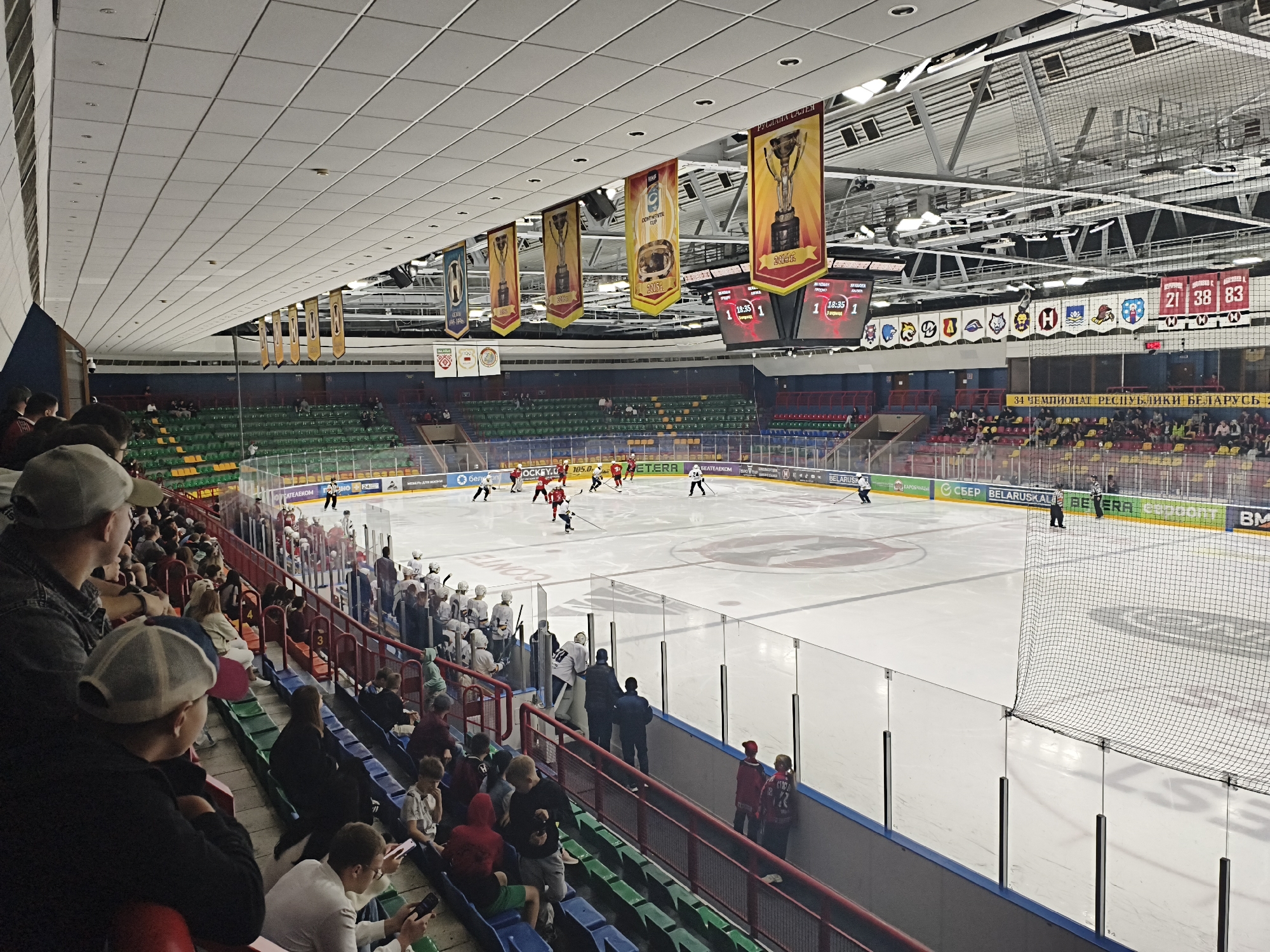 The image depicts an ice hockey arena with an ongoing game. Spectators are seated in green and yellow tiered seating, some in the foreground are looking towards the ice. The ice rink is visible with players in red and white uniforms engaged in the game. Banners with trophy graphics and team logos are suspended from the ceiling, along with scoreboard displays showing game times and scores. The arena appears to be the HC Neman ice rink in Hrodna, Belarus, given the prominent display of Belarusian league banners and sponsorships. The environment is indoor, with artificial lighting illuminating the scene.