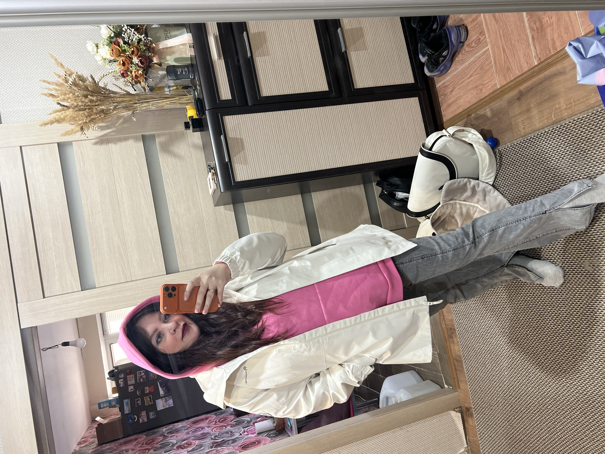 A woman is taking a selfie in a mirror inside a room. She is wearing a pink hoodie, a white jacket, and gray jeans. She is holding an orange smartphone. The room appears to be an apartment, with a refrigerator with magnets and pictures on it visible in the reflection. There is also a closet with light-colored doors, a cabinet with dark frames and light-colored panels, and various objects such as shoes, bags, and flowers scattered around. The floor is covered with a patterned rug. The mirror is framed, and the surrounding area is illuminated by artificial light. The location appears to be in an apartment, potentially in Noviy, Russia, although this cannot be confirmed with certainty from the image alone.