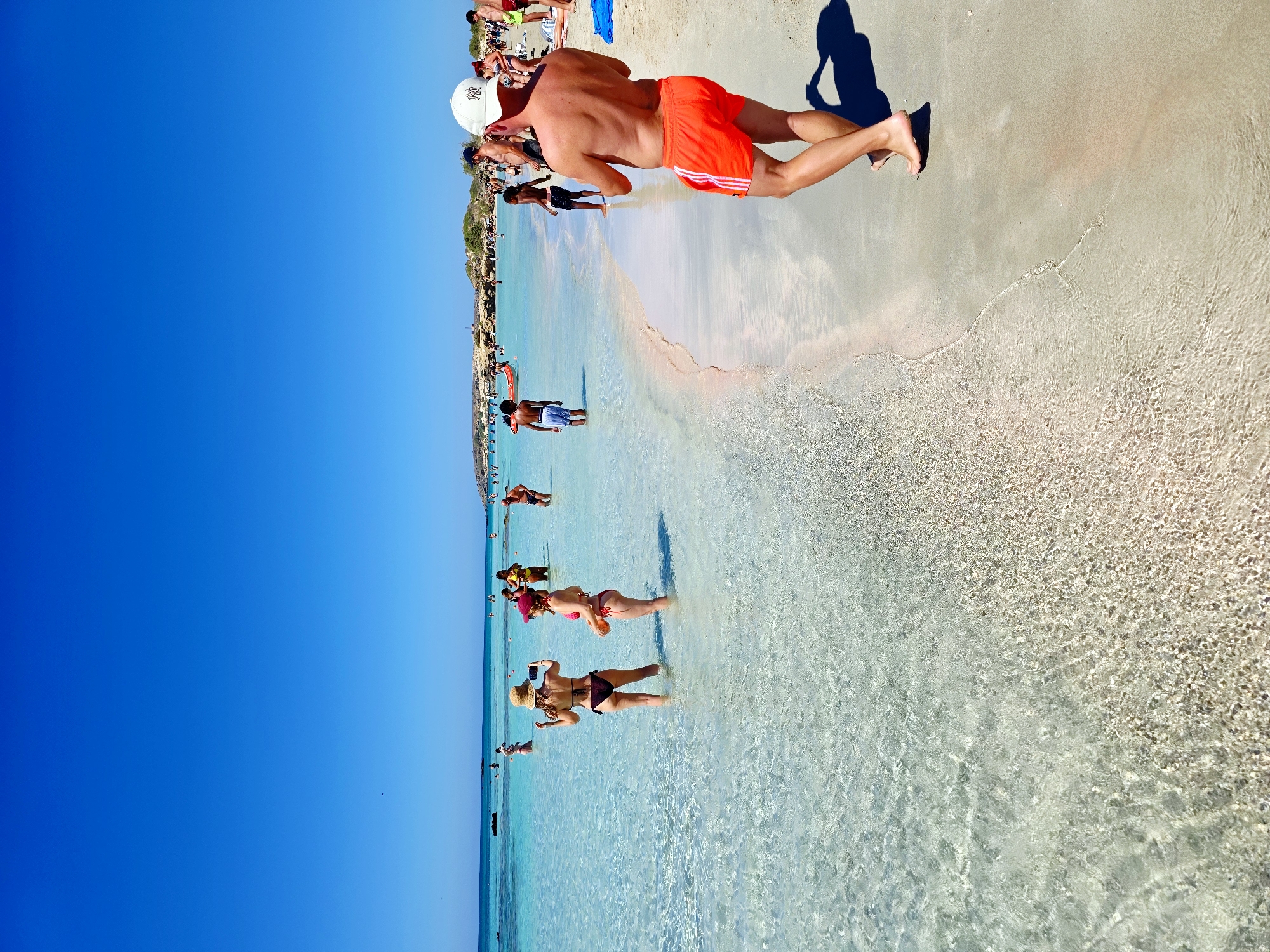 A clear, bright day at a beach in an unidentified location in Greece, possibly Nissi Beach, Ayia Napa, Cyprus, due to its distinctive turquoise waters. The foreground features a man in bright orange swimming trunks and a white baseball cap walking away from the camera, casting a shadow on the sand. The shallow, crystal-clear turquoise water extends to the left, revealing the sandy seabed. Several people are visible in the water and on the beach in the middle ground and background. Two women in bikinis are standing in the shallow water, one wearing a straw hat and holding a phone, the other holding an orange object. Other beachgoers, including children, are scattered across the scene, some wading, others interacting. The background shows a distant, rocky shoreline with sparse vegetation under a vivid, cloudless blue sky. The overall atmosphere is one of a sunny, active beach day with a focus on the clear waters and sandy shore.