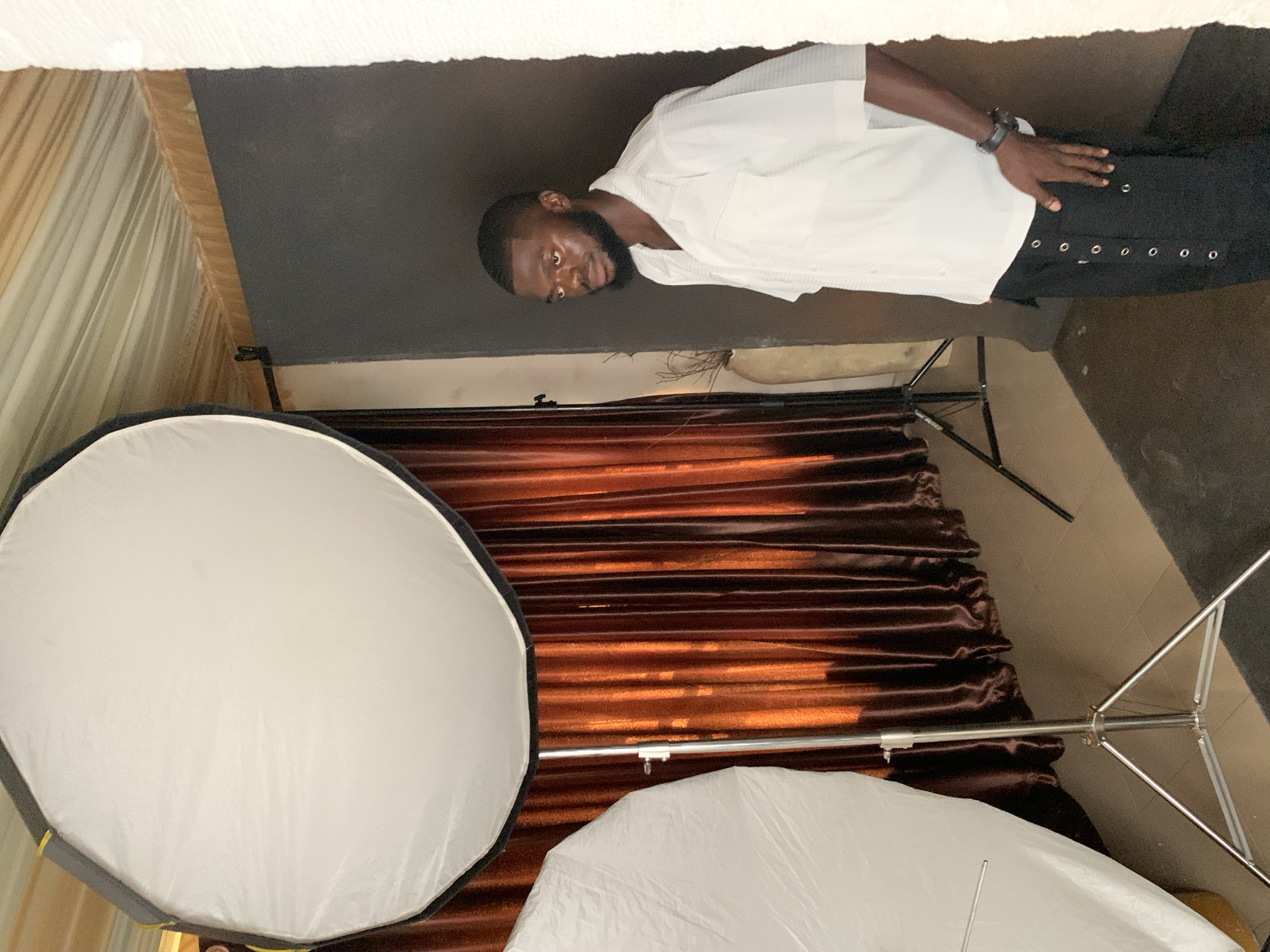 The media file features a young Black man in a studio setting, likely a photography studio, given the presence of studio lighting equipment. He is wearing a white short-sleeved collared shirt and black pants adorned with silver grommets. His left hand rests on his pants, and he wears a black wristwatch on his left wrist. The man has short black hair and a beard and is looking towards the camera with a neutral expression.
The background consists of a dark wall, brown satin curtains, and studio equipment. In the foreground, two large white softboxes are visible, indicating a controlled lighting setup. One softbox is octagonal, and the other appears to be umbrella-shaped. A metal stand for the lighting equipment is also visible. The overall impression is that of a posed portrait session. The lighting suggests it is indoors, and no external environmental cues like weather or time of day are discernible. The setting appears to be Gura Top, Nigeria, as provided by the location context. There is no visible text in the image.