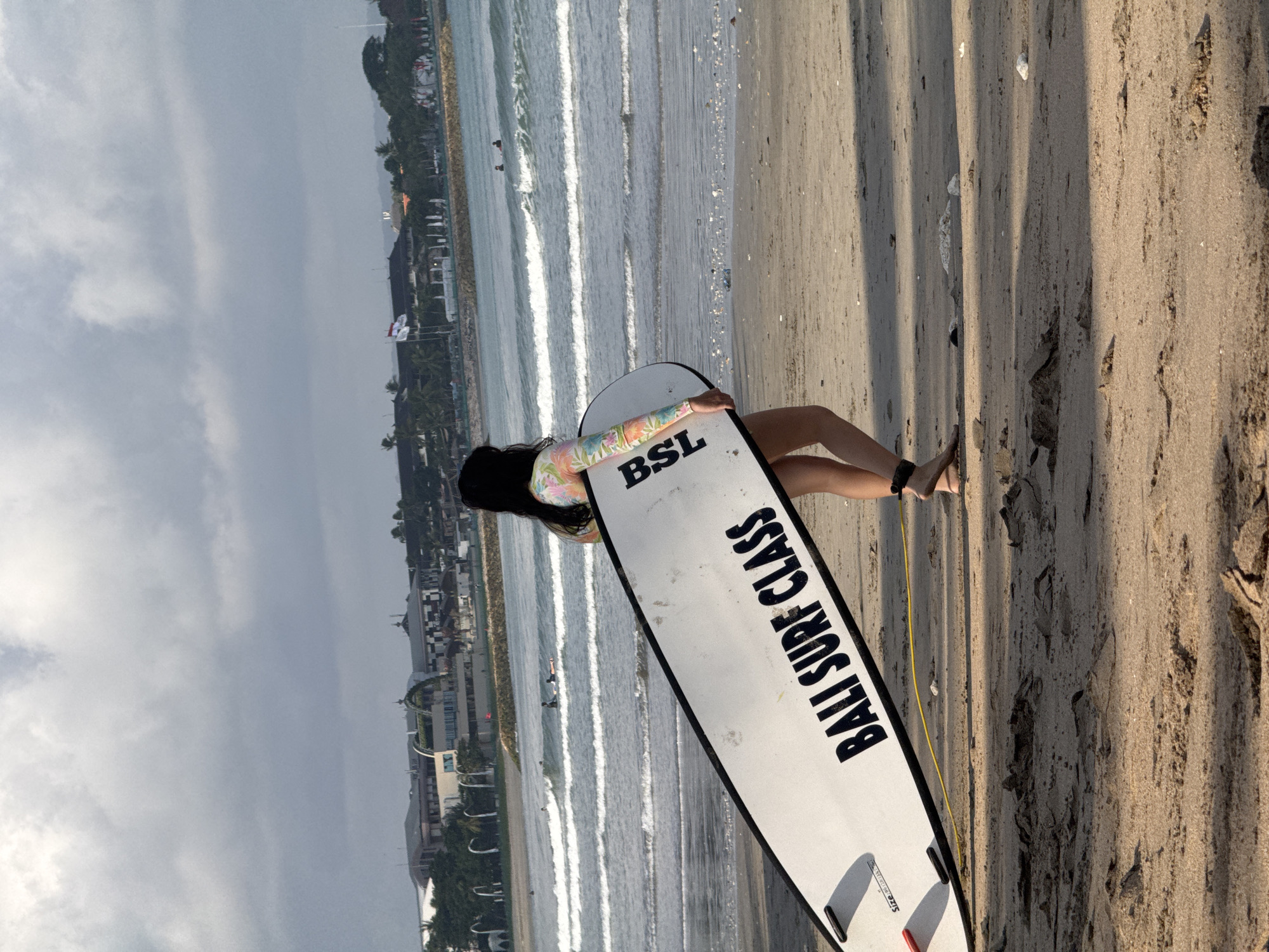 In Kuta, Indonesia, a woman stands on a sandy beach carrying a white surfboard. The surfboard has black lettering that reads "BALI SURF CLASS" and "BSL". She is wearing a long-sleeved rash guard with a colorful floral pattern and a black ankle leash. The ocean is to her left, with gentle waves and several surfers in the distance. Buildings and palm trees are visible in the background along the coastline. The sky is overcast with scattered clouds.