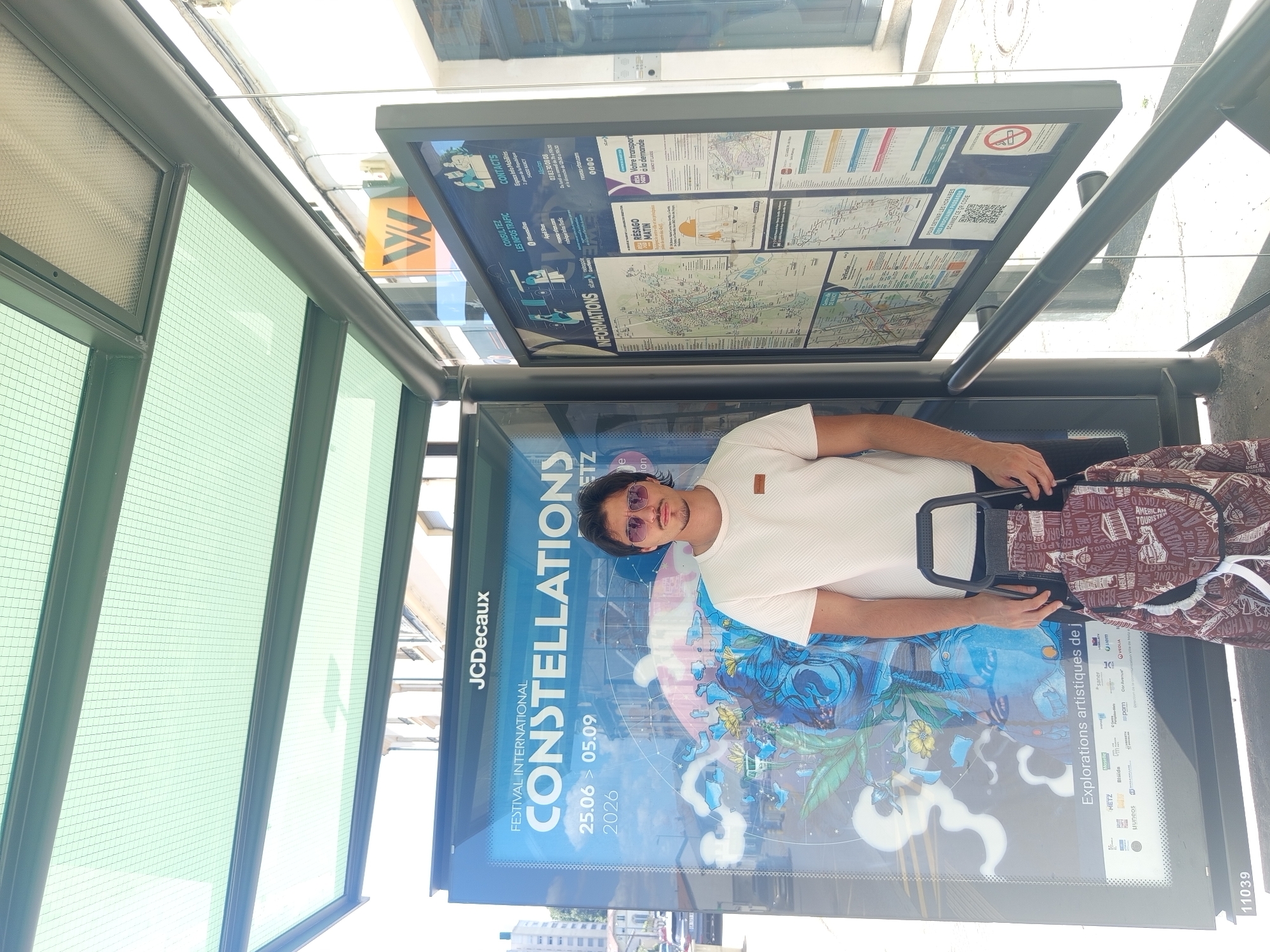 A young man with dark hair, sunglasses, and a light-colored t-shirt stands at a bus stop in Nancy, France. He is holding a patterned shopping trolley with a burgundy background and white text featuring various city names. Behind him is a bus stop shelter with a green, meshed roof. To his left is a large poster advertising the "FESTIVAL INTERNATIONAL CONSTELLATIONS" from June 25 to September 5, 2026, with the JCDecaux logo at the top. To his right, a large information board displays maps and details about local transportation services, including a QR code. The bus stop is situated on a street with buildings visible in the background.