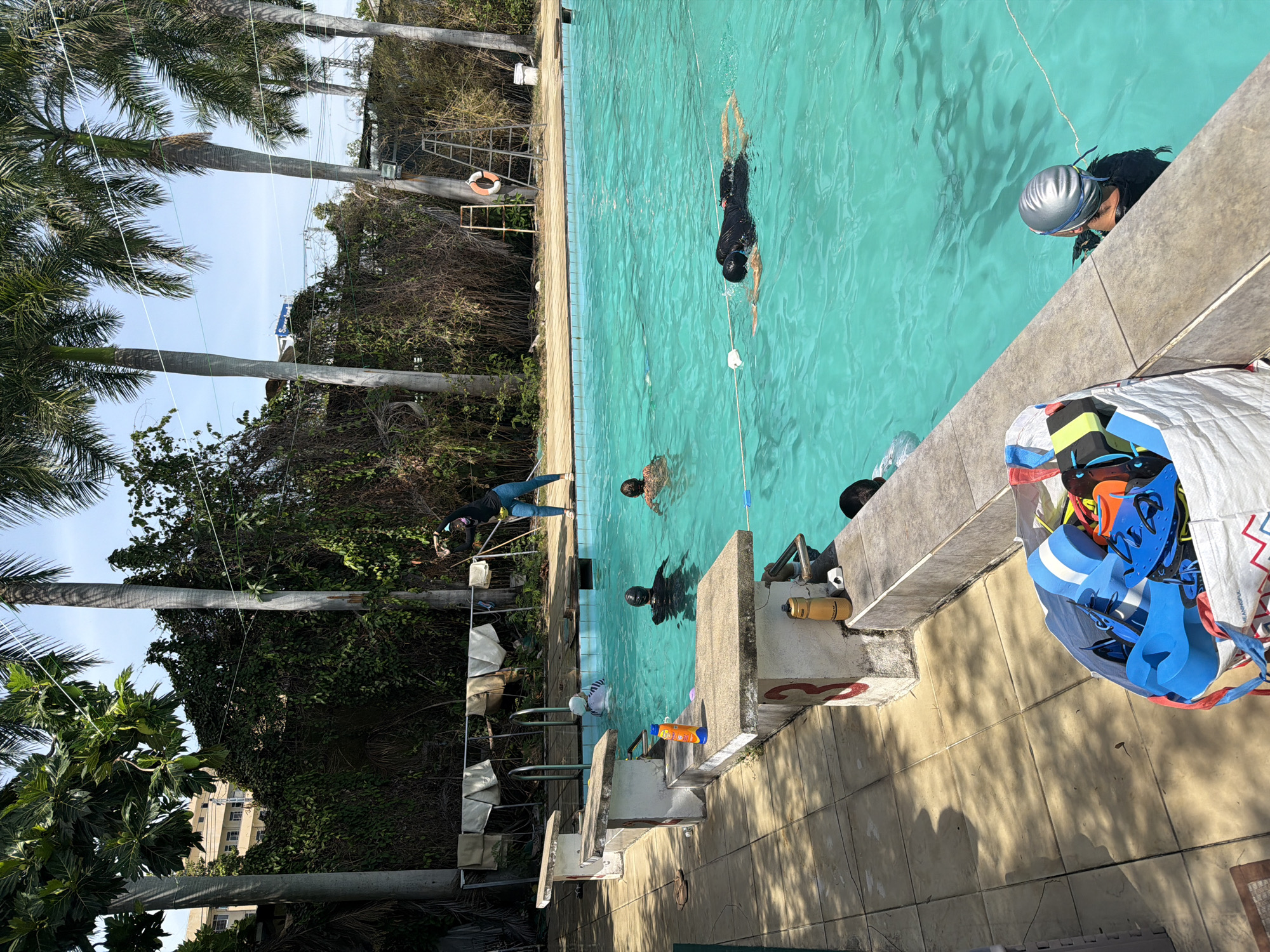 In Nha Trang Ward, Vietnam, a swimming pool area is depicted under a clear sky, with lush palm trees and dense foliage bordering the water. The pool itself is filled with vibrant turquoise water. Several individuals are engaged in swimming activities. One person, wearing a black swimsuit and cap, is performing a freestyle stroke. Another swimmer, also in a black swimsuit and cap, is visible further in the background, with only their head and shoulders above water. A third individual, wearing a white swim cap and goggles, is positioned near the diving boards. On the poolside, a diving board with the number "3" is visible, along with various swimming accessories in a large bag, including kickboards and pull buoys. A woman in athletic wear is standing at the edge of the pool, appearing to be instructing or participating in a warm-up. The environment includes a tiled walkway, a ladder leaning against a structure, and a life preserver ring.