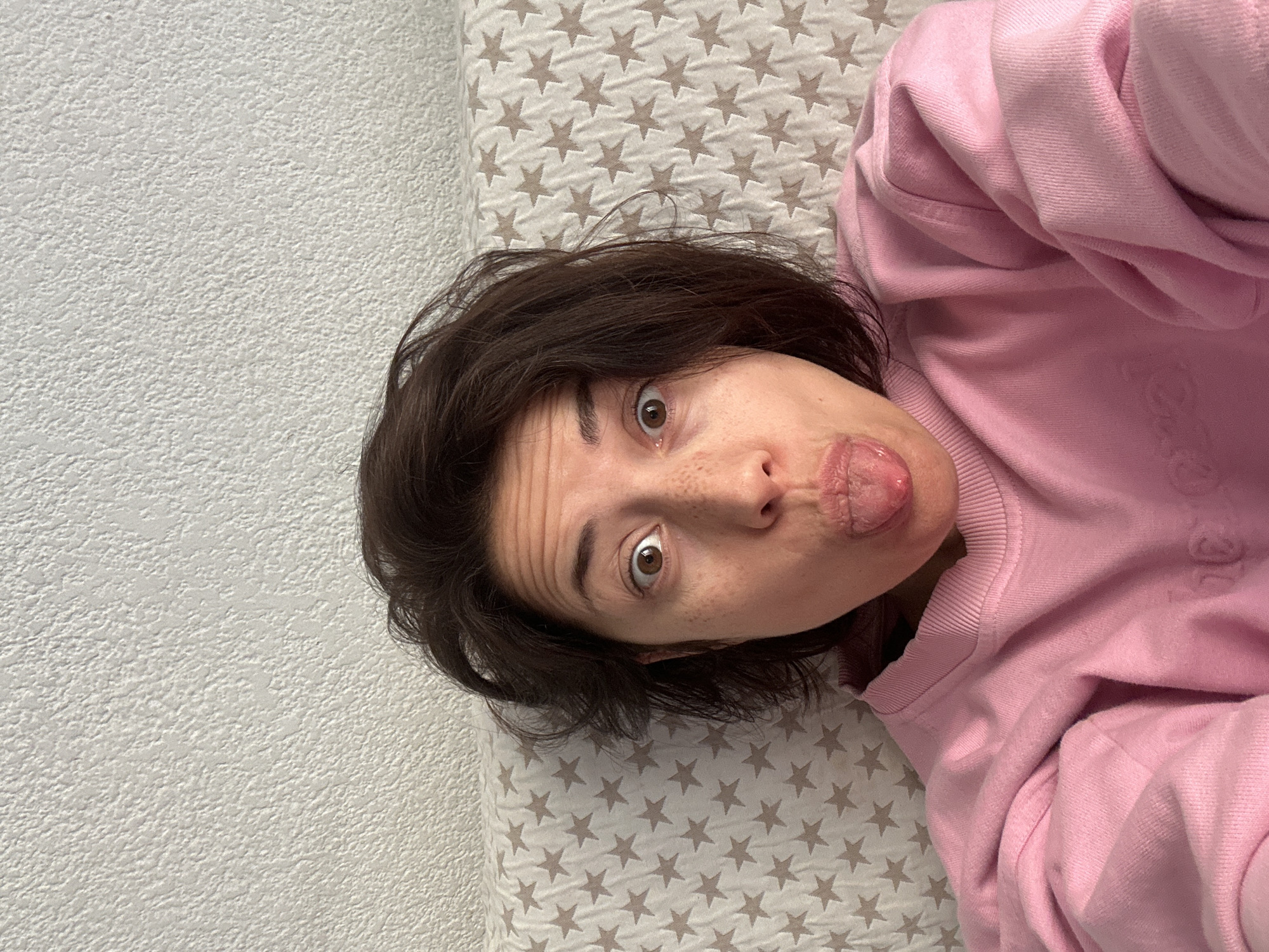 A portrait-oriented image captures a person making a playful expression while lying down indoors. The subject, an adult with short dark hair and brown eyes, looks directly at the camera with wide, expressive eyes and her tongue slightly extended. She is wearing a soft pink long-sleeved sweatshirt. The background consists of a light-colored, textured wall to the left and a light-toned fabric with a repeating pattern of brown stars to the right and beneath her head, suggesting a bedspread or blanket. The setting appears to be a brightly lit indoor space, such as a bedroom or living room. The overall mood is lighthearted and humorous. This scene is located in Barcelona, Spain. There is no visible text in the image.