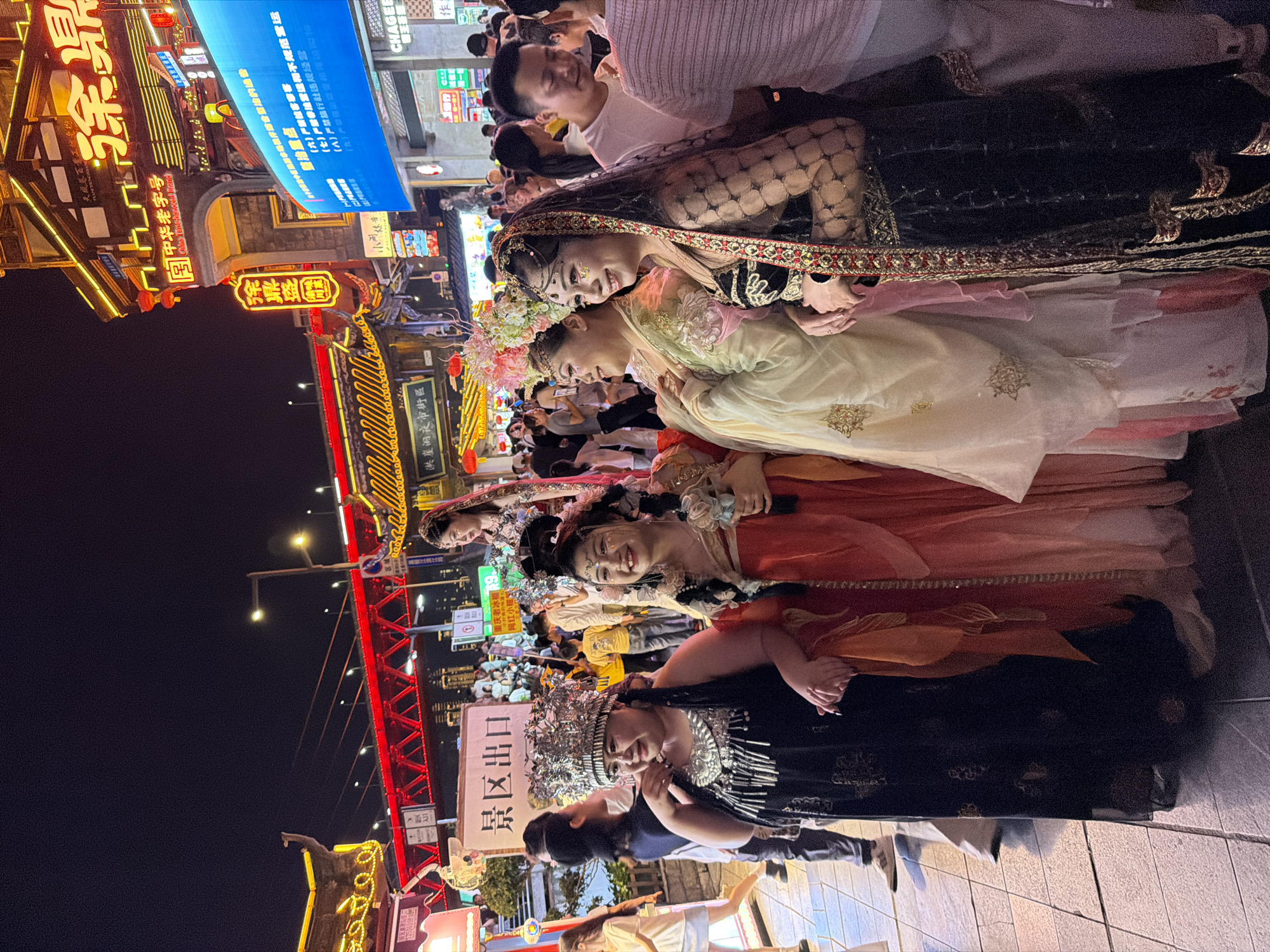 This media file captures a nighttime scene in the Yuzhong District of China, likely a bustling commercial area or night market. The primary subjects are several individuals dressed in elaborate, traditional-looking attire, reminiscent of cultural performances or historical reenactments. Two women in the foreground are the most prominent, adorned in ornate headpieces and flowing, richly colored garments. One wears a dark, intricately patterned dress with a jeweled headpiece and elaborate silver necklace, while another is dressed in lighter, pastel-colored robes with floral embellishments and a flower crown. Behind them, more people in similar attire are visible, suggesting a group or procession.
The setting is characterized by vibrant neon signs and illuminated storefronts, typical of a lively urban nightscape. Red architectural elements, possibly part of a bridge or building facade, are visible, adding to the festive atmosphere. The ground appears to be paved with stone tiles. The activity seems to be a public gathering or event, with a crowd of onlookers visible in the background, some of whom are taking pictures. The overall mood appears celebratory and engaging, with the subjects in the foreground smiling and interacting.
Visible text includes Chinese characters and some English. Notably, a sign reads "景区出口" (Scenic Area Exit). Another sign displays "洪崖洞夜市街区" (Hongyadong Night Market Street Area), providing a specific location clue. There are also regulatory notices with headings like "整治重点" (Rectification Key Points) and enumerations of prohibited activities. The presence of a "China Time-honored Brand" sign also indicates a place with historical significance or traditional businesses. The time of day is clearly night, as evidenced by the artificial lighting and the dark sky.