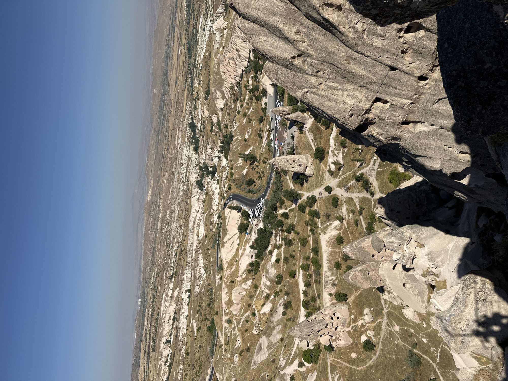 An elevated, wide-angle shot captures the unique geological formations of Cappadocia, a region in Uçhisar Beldesi, Turkey. The foreground is dominated by the rough, textured surfaces of large rock formations, casting prominent shadows onto the terrain below. A network of pathways winds through the valley floor, dotted with numerous "fairy chimney" rock structures, many of which exhibit carved openings, suggesting ancient human habitation or use. Lush green trees are scattered amongst the rocky landscape, contrasting with the predominantly tan and white hues of the eroded earth. A paved road curves through the scene, bordered by a parking area with several white vehicles. In the mid-ground, more of these distinctive rock formations are visible, alongside a scattering of buildings. The distant background reveals a vast, hazy landscape of rolling hills and a clear, bright blue sky. The overall scene depicts a natural wonder with evidence of human interaction.