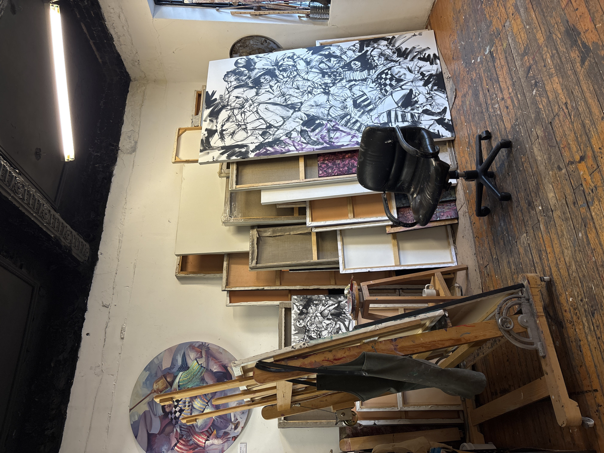 The image depicts an artist's studio, seemingly in a state of organized chaos. The room has hardwood flooring and white walls, with a black area likely containing a doorway or large window frame on one side. A fluorescent light fixture illuminates the left wall. Various paintings in different sizes and stages of completion lean against the wall, with a large black and white abstract piece dominating the top. A black office chair is positioned near these canvases. An art easel is situated at the bottom of the frame, covered with what looks like an apron or cloth stained with paint. A round painting with abstract shapes and figures is visible towards the lower left. The location is given as the city of Unknown, Unknown.