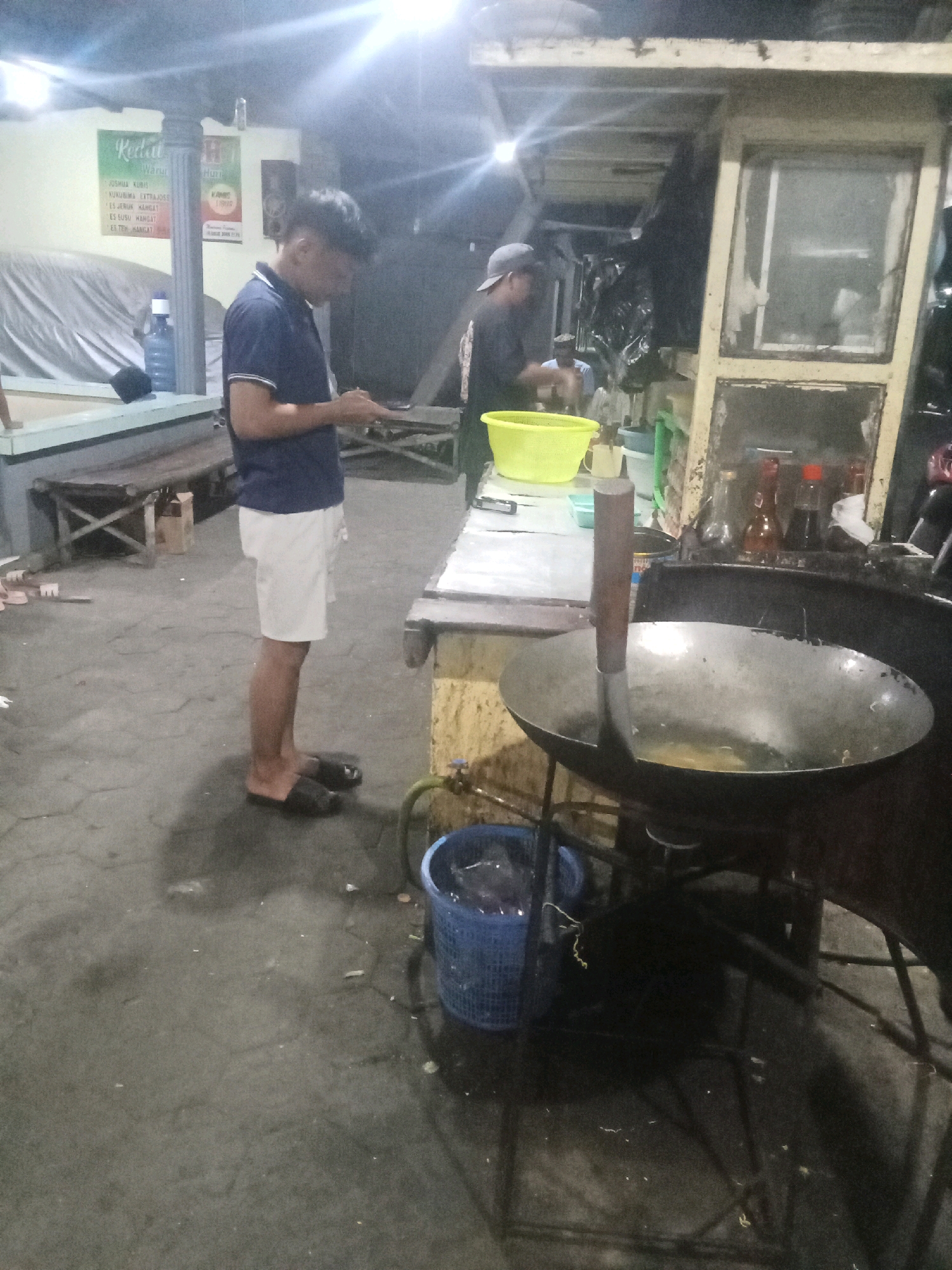 The image depicts an outdoor food stall setting in the city of Ngumpul, Indonesia, likely at night due to the artificial lighting. In the foreground, a man wearing a dark blue polo shirt and white shorts stands facing to his left, holding a mobile phone. He is wearing black flip-flops. Behind him, another person, partially obscured, wearing a dark shirt and a baseball cap, appears to be working at the stall.
The stall itself is characterized by a wok being heated, with oil inside, and a large metal spatula resting in it. Various items are visible on the stall counter, including bottles of sauces, a yellow plastic basin, and what appear to be plastic containers. To the left of the stall, a large blue plastic basket filled with water and some items is visible on the ground. The ground surface is paved with hexagonal tiles. In the background, there are tables and chairs, and a large white tarp. A sign with text in Indonesian, including "Kedai Warung" and menu items, is mounted on the wall behind the man in the foreground. The overall environment suggests a casual, operational food vendor.