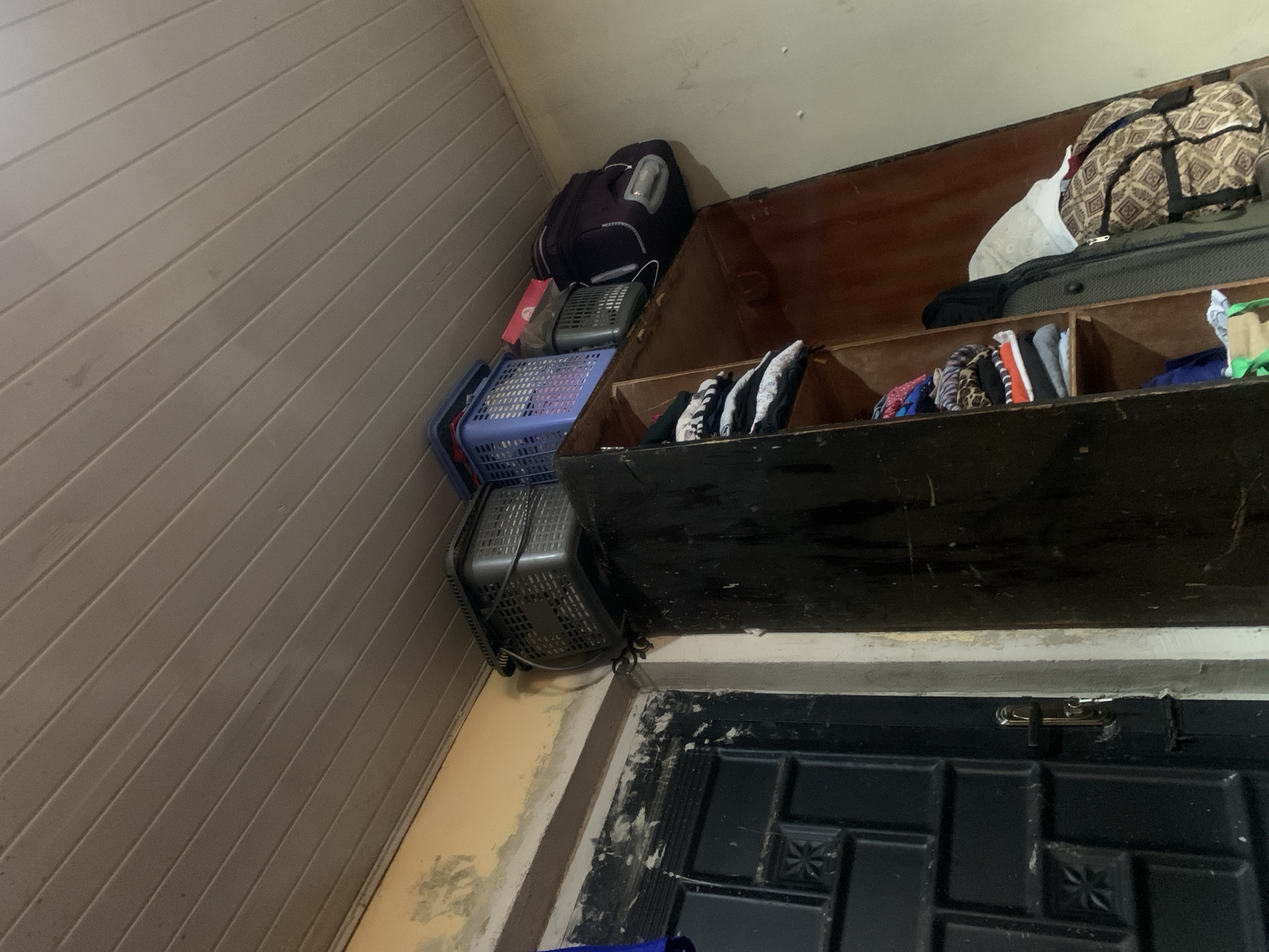 The image shows the top of a wooden wardrobe with several items stored on top. There are three baskets, two dark-colored and one blue, stacked on top of the wardrobe. A large dark purple backpack with white accents is also on top, leaning against the wall. Inside the wardrobe, on a shelf, are stacks of folded clothing, predominantly dark and white patterned items. To the right of the wardrobe, another shelf contains a suitcase and a patterned bag. The background shows a textured wall and a portion of a dark door with a metallic handle and a key in the lock. The ceiling is made of light-colored wooden planks.