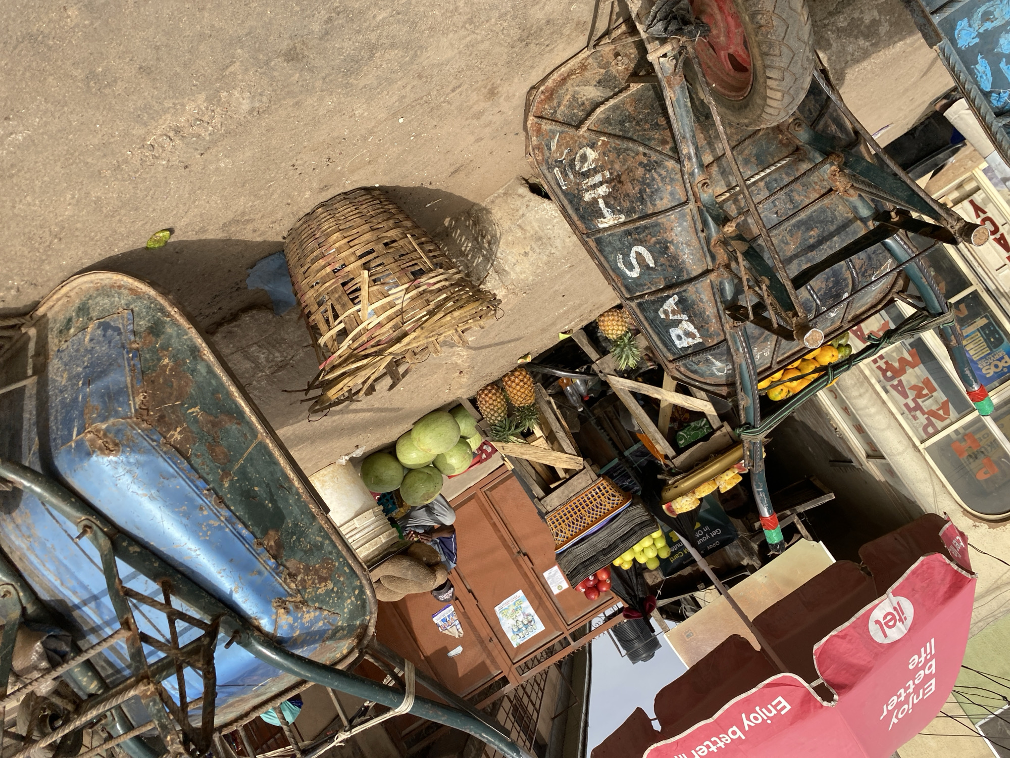 This media file presents a vibrant outdoor market scene in Bukuru, Nigeria, captured during daytime under bright sunlight. The ground is a mix of concrete and dirt, with strong shadows indicating the time of day.
The foreground is dominated by two well-used, rustic carts, characteristic of street vendors. The cart on the left is predominantly blue and appears to be a wheelbarrow-style carrier. The other cart, on the right, is dark, metallic, and exhibits significant rust, featuring a visible red wheel and a frame wrapped with rope. The side of this cart is painted with white text, clearly reading "BASHID" and partially "S HI", likely part of a name or descriptor.
Between the carts, a large, intricately woven bamboo basket rests on the ground. A diverse array of fresh fruits is displayed for sale on a wooden stand or directly on the carts. These include several green watermelons, multiple pineapples, and piles of yellow-green fruits (possibly oranges or apples) and some red fruits. A person is partially visible, seated behind the fruit display, near a small, brown kiosk or stall.
A large red umbrella, providing shade, stands prominently in the scene. It features the "itel" logo and the slogan "Enjoy better life," with a portion of the text "Enjoy bette" also visible. Other signs and posters are present, advertising various services; visible text includes "POS SERVICES," "Opay," "Get your in ON," "M Card," and "nute," suggesting mobile money and communication services are accessible in the area. The overall impression is a bustling marketplace reflecting daily commerce.
