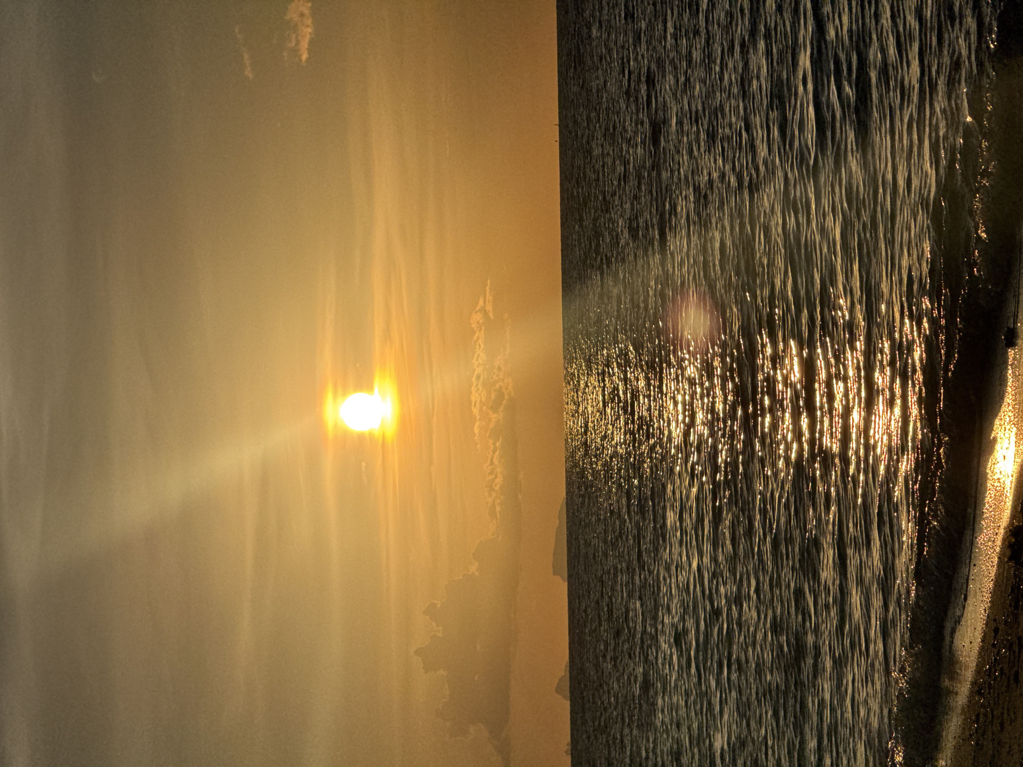 This media file depicts a sunset at Baan Tai, Thailand. The image is split vertically, with the left side showing the sun setting behind a hazy, golden sky. Streaks of light emanate from the sun, creating a diffused glow that illuminates wispy clouds. On the right side of the image, the ocean is visible, with the setting sun casting shimmering reflections on the rippling water. The waves appear dark, contrasted by the bright glints of sunlight. The edge of the land, possibly a beach, is visible on the far right, with wet sand reflecting the golden light. The overall mood is serene and peaceful, capturing the beauty of a tropical sunset. The time of day is clearly sunset, and the weather appears clear with some atmospheric haze. There is no visible text in the image.