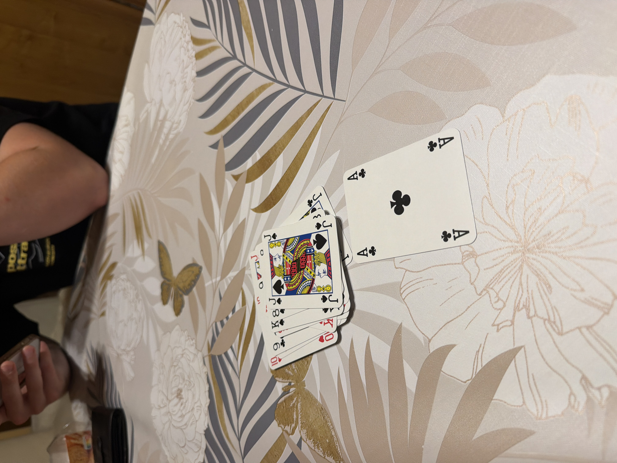 The image depicts a tabletop scene with playing cards. A hand is visible on the left edge, holding more cards. On the table, there is a pile of cards with the Jack of Spades on top, followed by the 8 of Hearts, 10 of Hearts, and others. Next to this pile is a single card, the Ace of Clubs. The table has a patterned tablecloth with floral and leaf designs in shades of beige, gray, and gold. The environment is domestic, possibly in Burgas, Bulgaria, but there are no explicit indicators to confirm the city.
