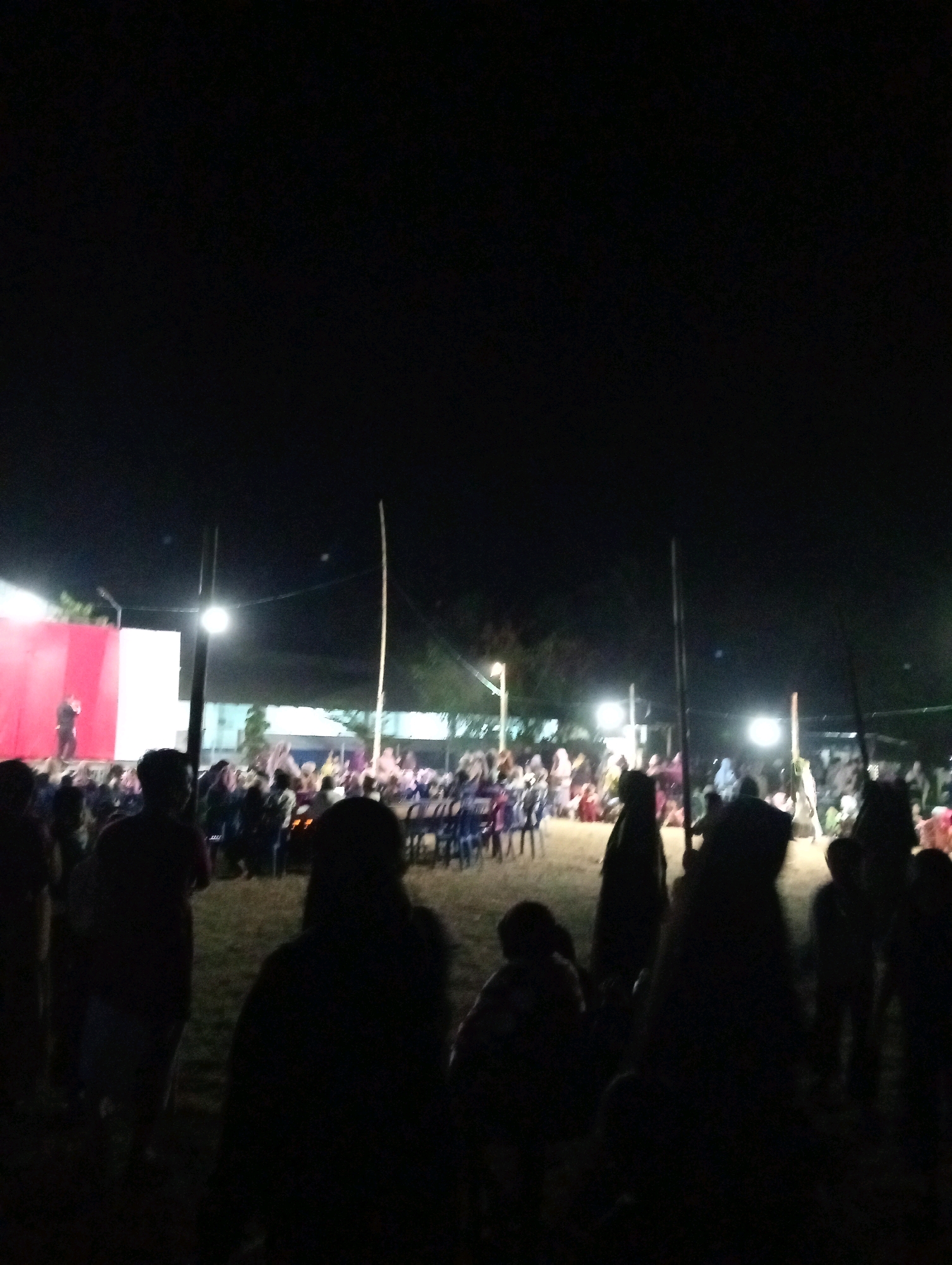 This is a nighttime outdoor photograph taken at an event in Pagerungan Kecil, Indonesia. A stage is set up on the left side of the image, with a large red and white backdrop. A single person, likely a performer, is standing on the stage and appears to be speaking or singing into a microphone. In front of the stage, a crowd of people is gathered, seated on blue chairs. The scene is illuminated by bright, artificial lights, casting a warm glow on the area. The foreground of the image is dominated by silhouettes of people standing and watching the event, their features indistinct in the low light. The overall impression is of a community gathering or performance taking place after dark.