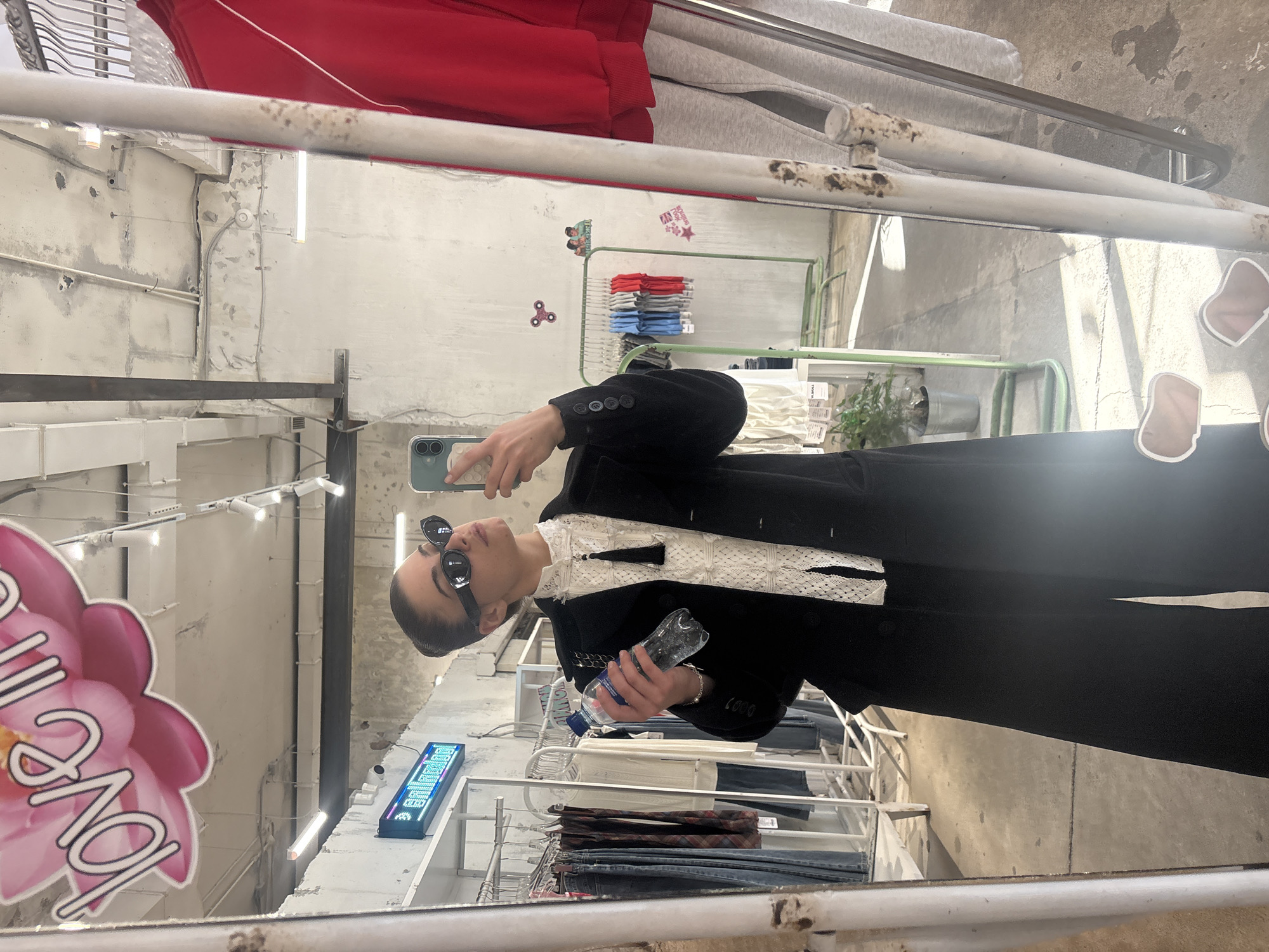 A person takes a mirror selfie while in a store. The store has a clothing rack with folded jeans on the bottom shelf and t-shirts on the top rack. The walls of the store appear to be concrete. There is a pink flower decal on the mirror. The person is wearing a black blazer and sunglasses, and is holding a water bottle. The reflection also shows a sign on the wall and other clothing racks. The location is in a store in Moscow, Russia.