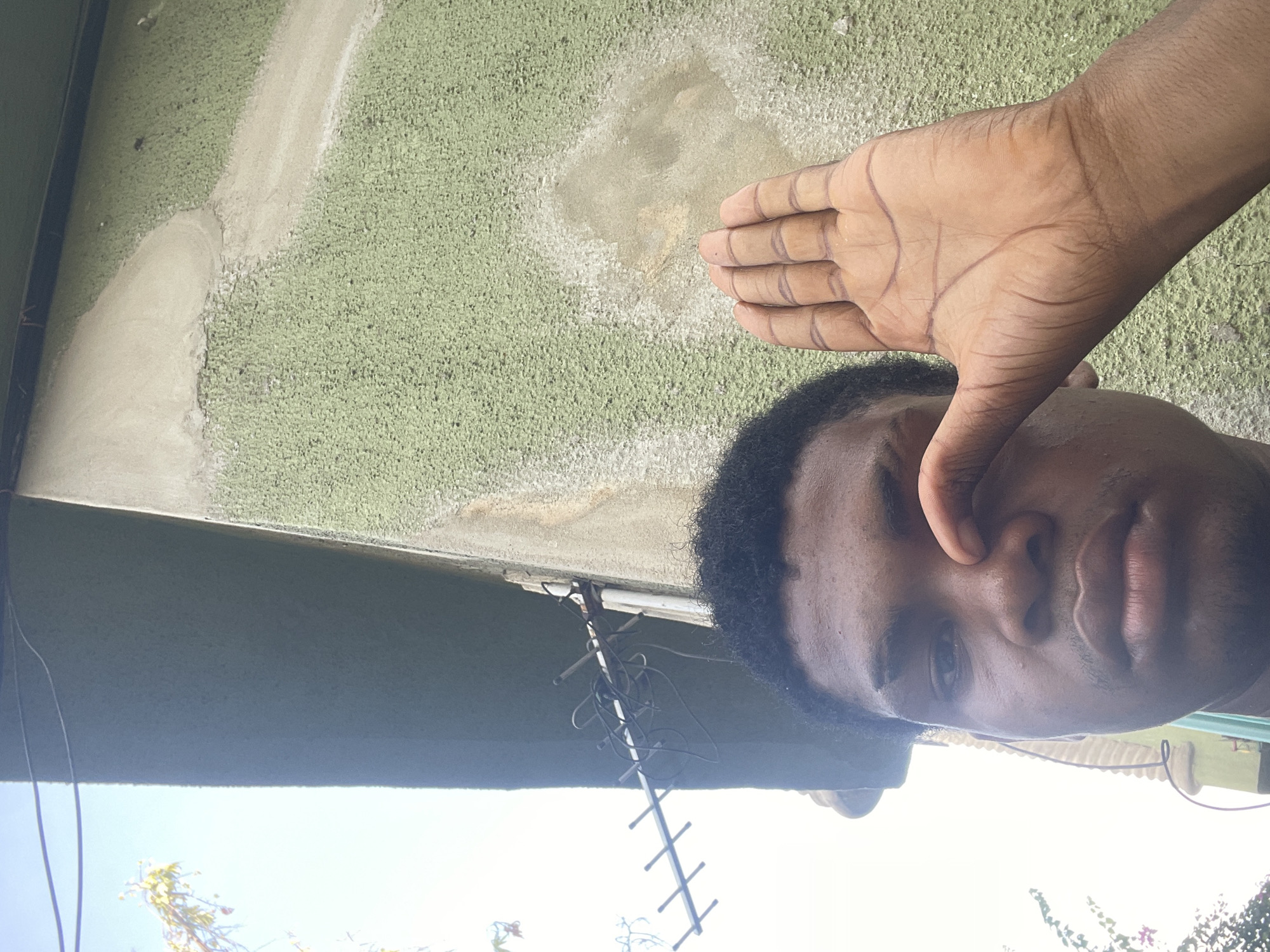 A selfie captures a person with dark hair and skin, their right hand raised, fingers extended, nearly obscuring the right eye. The subject appears to be outdoors, and a portion of a green, textured wall with irregular beige patches forms the background. An antenna extends from under the wall, suggesting a building. The location is possibly in Ado, Nigeria, based on the user's request, but there is no concrete visual evidence within the image to verify this.