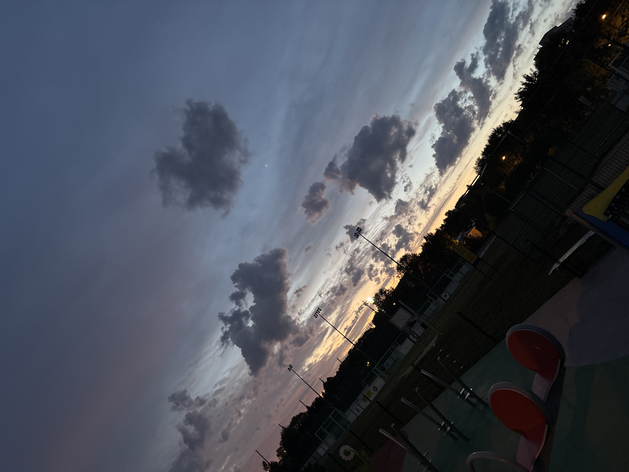 The image depicts an outdoor scene at dusk, featuring a sky filled with dramatic clouds tinged with sunset colors. In the foreground, playground equipment is visible, including two red seats on a see-saw-like structure. The middle ground shows a fenced area, possibly a sports field or court, illuminated by tall lampposts. Trees line the perimeter of this area, and some buildings are visible in the background. The overall impression is that of a public park or recreational facility in Lublin, Poland, as the sun sets, casting long shadows and creating a moody atmosphere.