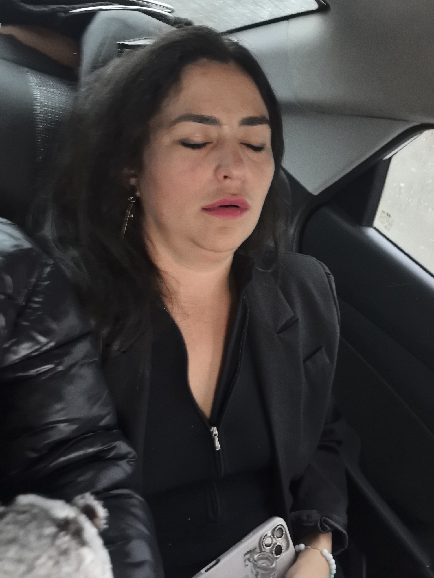 A woman with dark hair and wearing dark clothing sits in the back seat of a car. Her eyes are closed and her head is slightly tilted back, suggesting she may be asleep. She is wearing an earring and a bracelet. She is holding a cell phone. Rain is visible on the car window. It is not possible to confirm from this image that she is in Bolgury, Russia.