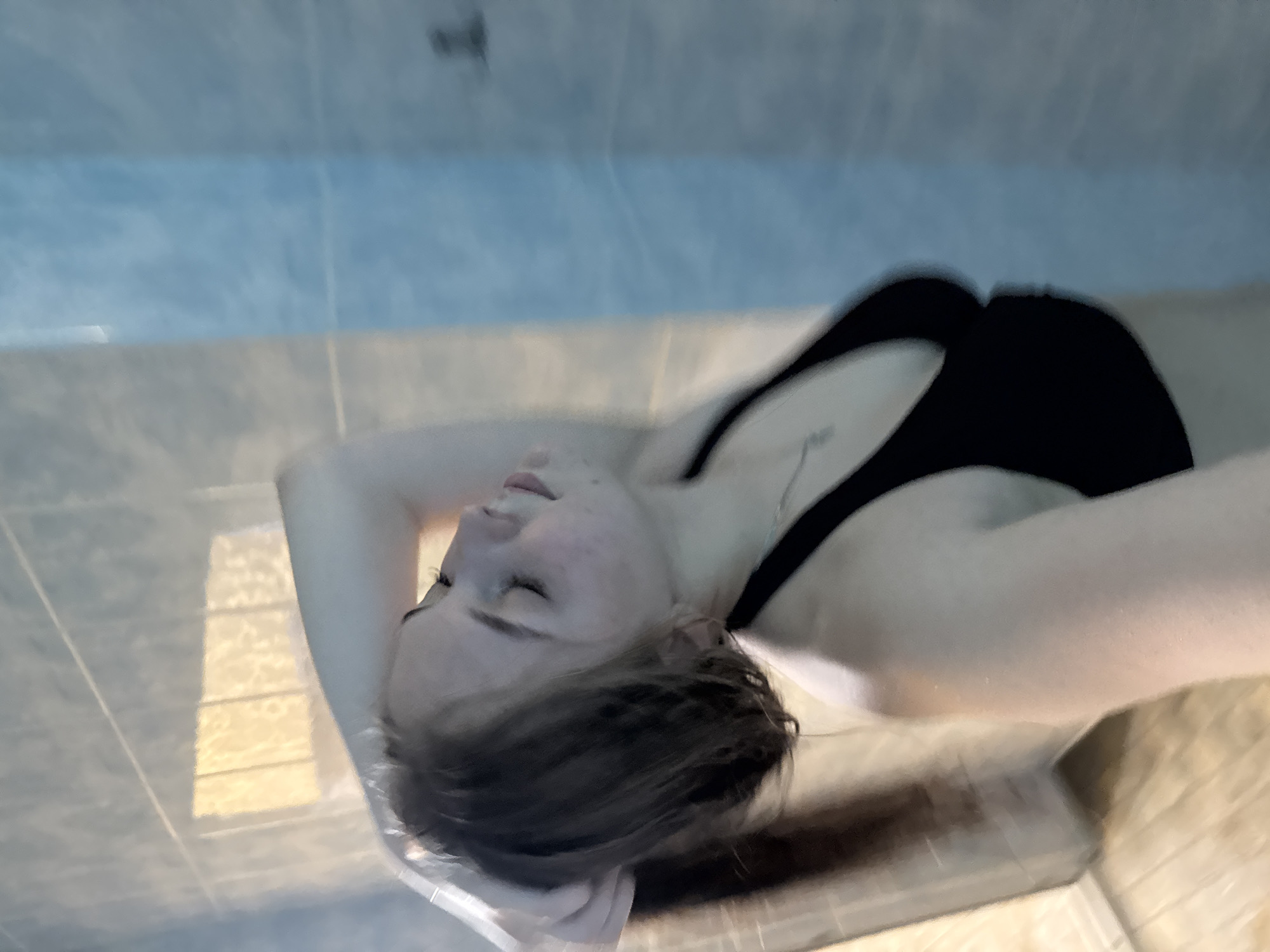 A woman is floating in a sensory deprivation tank. She has brown hair and fair skin. She is wearing a black swimsuit. Her eyes are closed, and her face is relaxed. Her hands are resting behind her head. The tank is lined with blue and white tiles. This facility may be located in the city of Chaikovsky, Russia.