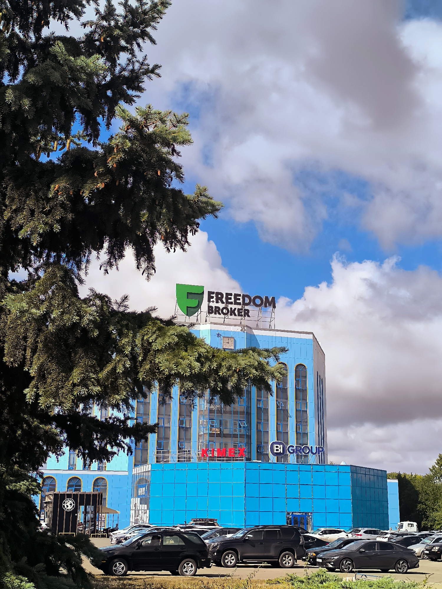 This image depicts a modern, blue-colored building with prominent signage for "FREEDOM BROKER" and "BI GROUP" on its upper levels. The building appears to be a commercial or office complex. In front of the building, a parking lot is filled with a variety of cars. In the foreground, a large, dark green coniferous tree partially obscures the view of the building and the parking lot. The sky is a mix of blue with scattered white and gray clouds. The scene appears to be set in an urban environment, specifically the city of Qarağandı city administration, Kazakhstan. No people are clearly visible in the image, and the focus is on the architecture and the surrounding environment.