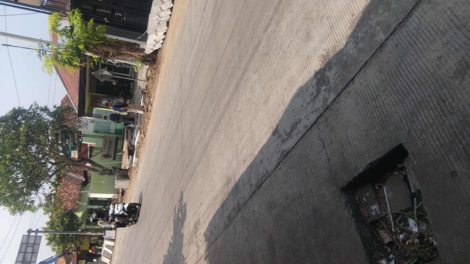 The image depicts a street scene in Batang, Indonesia, likely on a sunny day. In the foreground, a cracked concrete sidewalk with a hole filled with debris is visible. The road surface is asphalt with visible tire marks and shadows cast by trees.
On the road, a man wearing a helmet is riding a white motorcycle with another person also wearing a helmet as a passenger. They are traveling from left to right across the frame.
In the background, there are several buildings with red tile roofs. One building has a green facade and a sign that reads "APOTEK BENMARI". Two men are standing near this building. One man is wearing a dark t-shirt and shorts, and the other is wearing a light-colored sarong and a dark t-shirt. Further down the street, there are more buildings, some with open doorways. A black scooter is parked near one of the buildings. On the right side of the image, a stack of concrete blocks is visible. Trees with green foliage are present throughout the scene, casting shadows on the road. The sky is clear and bright.