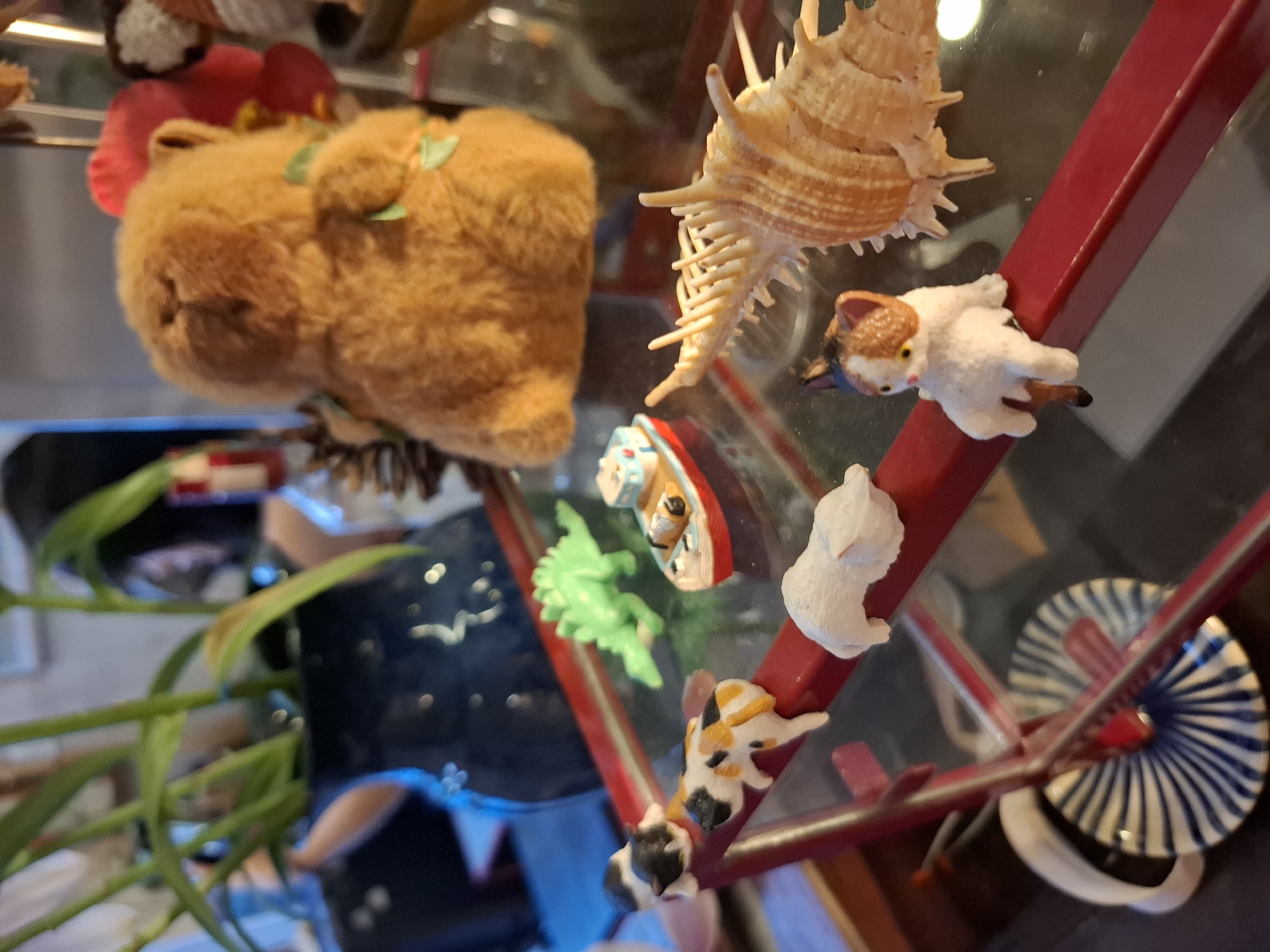 This is a close-up, eye-level shot of a glass shelf unit with various miniature decorative items. The shelf unit is red and appears to be part of a tiered display. On the shelf, there is a fuzzy orange teddy bear sitting behind a small green plastic dinosaur and a red and white toy boat. To the left of the dinosaur, a row of three small figurines are perched on the edge of the shelf: a black and white cat, a calico cat, and a white cat. To the right of the toy boat is a large, spiky, tan-colored seashell. Further to the right and slightly behind the seashell, the blurred outline of another decorative item is visible. In the background, a dark blue vase filled with green plants is partially visible on a higher shelf, along with other blurred objects. The lighting is somewhat dim, with some reflections on the glass surfaces. The image does not provide any information or visual cues to determine the specific location within Nha Trang Ward, Vietnam.