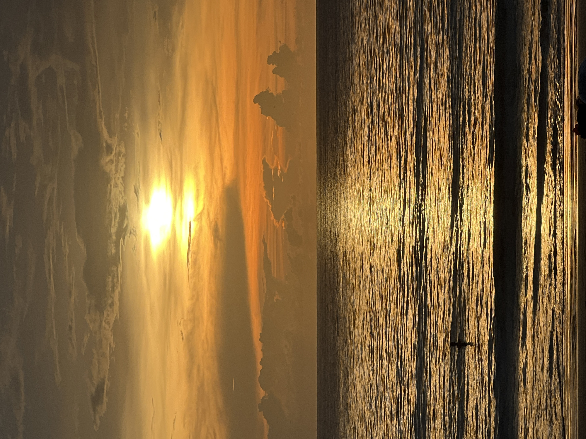 A vertical photograph captures a sunset over the ocean, likely in an unspecified location. The left side of the image depicts the sky filled with clouds and a bright sun. The right side shows the ocean with the sun's reflection creating a shimmering, golden effect. A small boat is visible in the water near the center-bottom of the frame.
