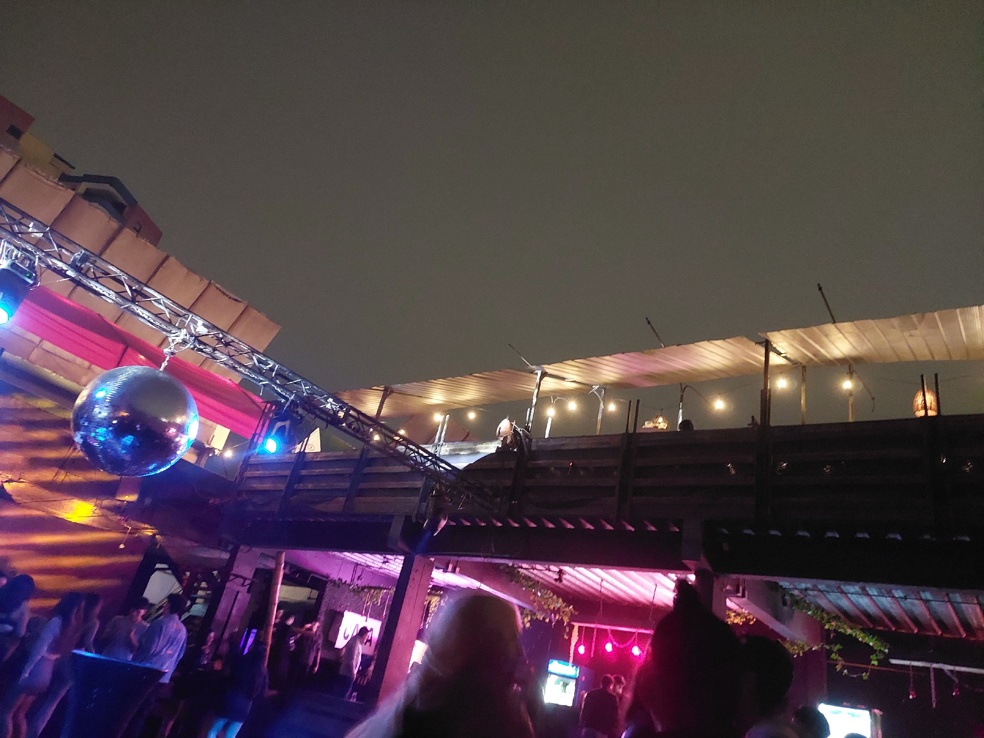 The image shows a crowded outdoor party in Arica, Chile, at night. A large disco ball hangs from a metal structure with lighting, casting reflections on nearby surfaces. People stand and mingle under the dimly lit structure and a higher wooden deck decorated with string lights. Pink and purple lighting illuminates the area. The sky is overcast.