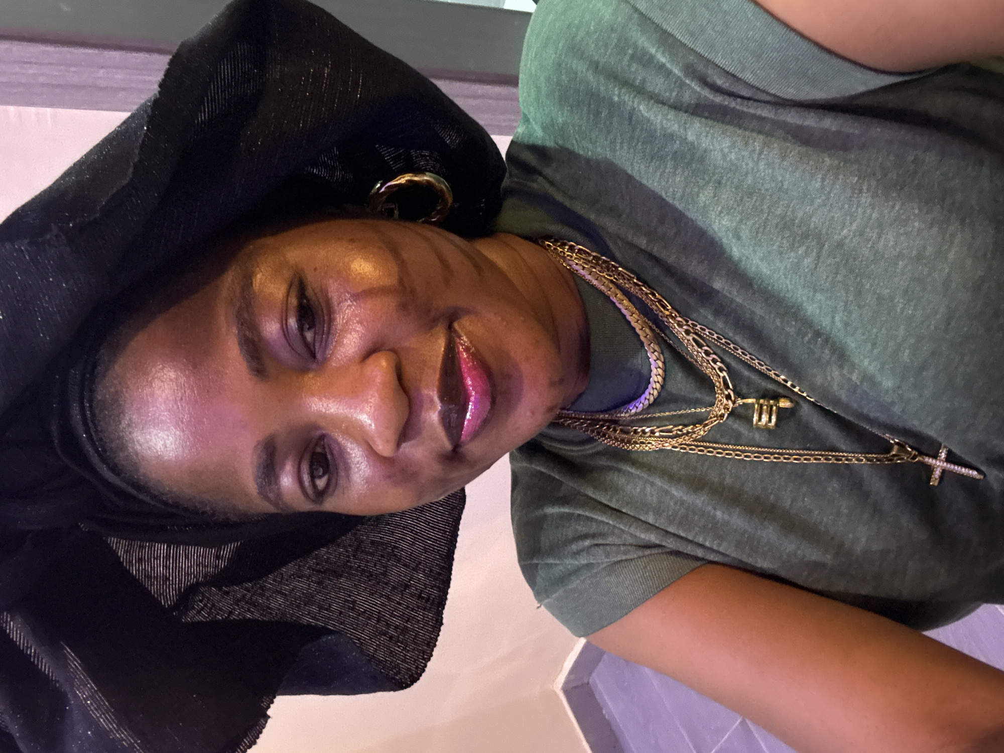 A medium close-up shot shows a woman wearing a black headwrap and a green t-shirt. She has on multiple gold necklaces, one with a cross pendant. She is wearing hoop earrings and has on pink lipstick. The background appears to be an indoor setting with neutral-colored walls and a tiled floor. The lighting is dim, casting shadows on her face. The image is taken from a slightly upward angle. The woman's facial features are clearly visible, and she appears to be looking directly at the camera. The overall impression is that of a selfie. There is no indication of the city of Ajara, Nigeria, within the image itself.