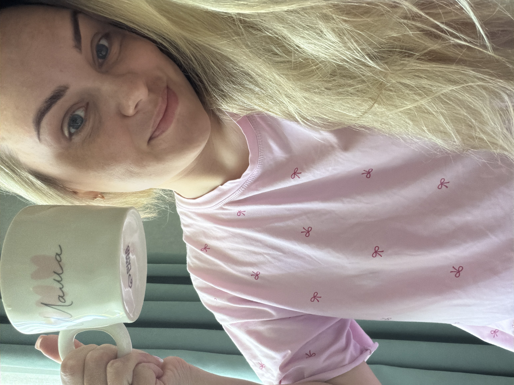 A selfie shows a fair-skinned woman with blonde hair smiling and holding a mug that says "Mama" in cursive. She is wearing a pink shirt with a pattern of small red bows. The background appears to be an interior space, possibly a home, with a light green surface visible behind her arm. The exact location in Votkinsk, Russia, is not discernible from the image.