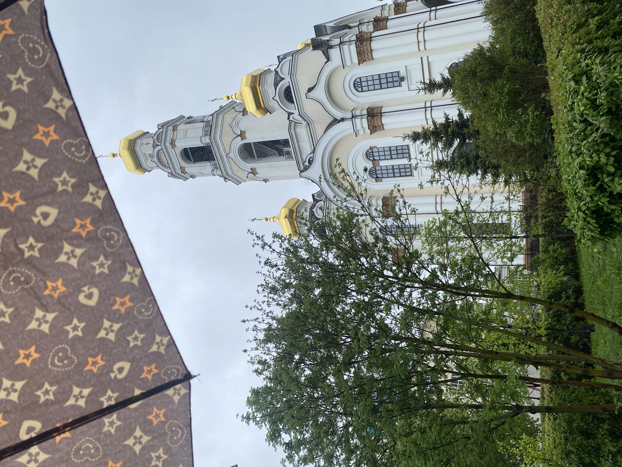 The image captures a view of the Alexander Nevsky Cathedral in Yekaterinburg, Russia, partially obscured by a tree and an umbrella. The cathedral is a white and cream-colored building with golden domes. The sky is overcast. A brown umbrella with a patterned print of shapes like hearts, stars, and other symbols occupies the lower left corner of the frame. A tree with green leaves stands in the foreground, partially obscuring the view of the cathedral. Green bushes are visible on the right side of the frame.