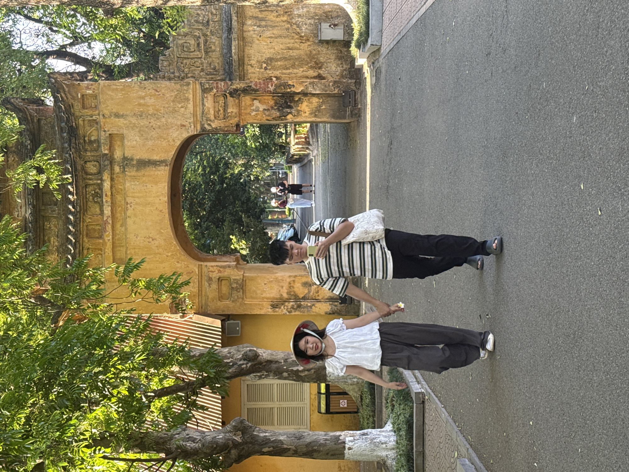 The image shows a couple holding hands on a street lined with trees and old buildings in Hà Nội, Vietnam. The man is wearing a striped shirt and holding a phone. The woman is wearing a white shirt and a Vietnamese conical hat. They are walking in front of an old archway with intricate carvings. Other people are visible in the background, walking under the archway. The street is paved with asphalt. A tree with green foliage is visible on the left side of the image. A building with a window and a striped awning is also visible.
