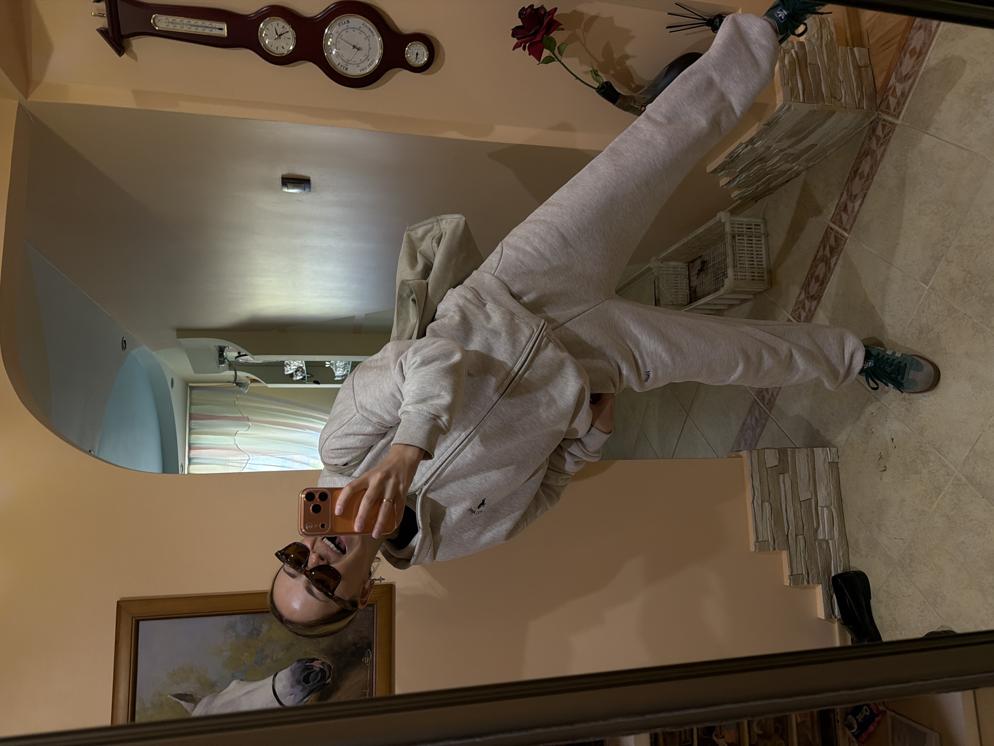 The image is a selfie taken in a mirror. The subject is wearing sunglasses and a light-colored tracksuit, holding a smartphone to capture their reflection. The background includes a hallway with peach-colored walls and a tiled floor. There is a painting on the wall, a clock, and a flower arrangement. The location is Kamyshin, Russia.