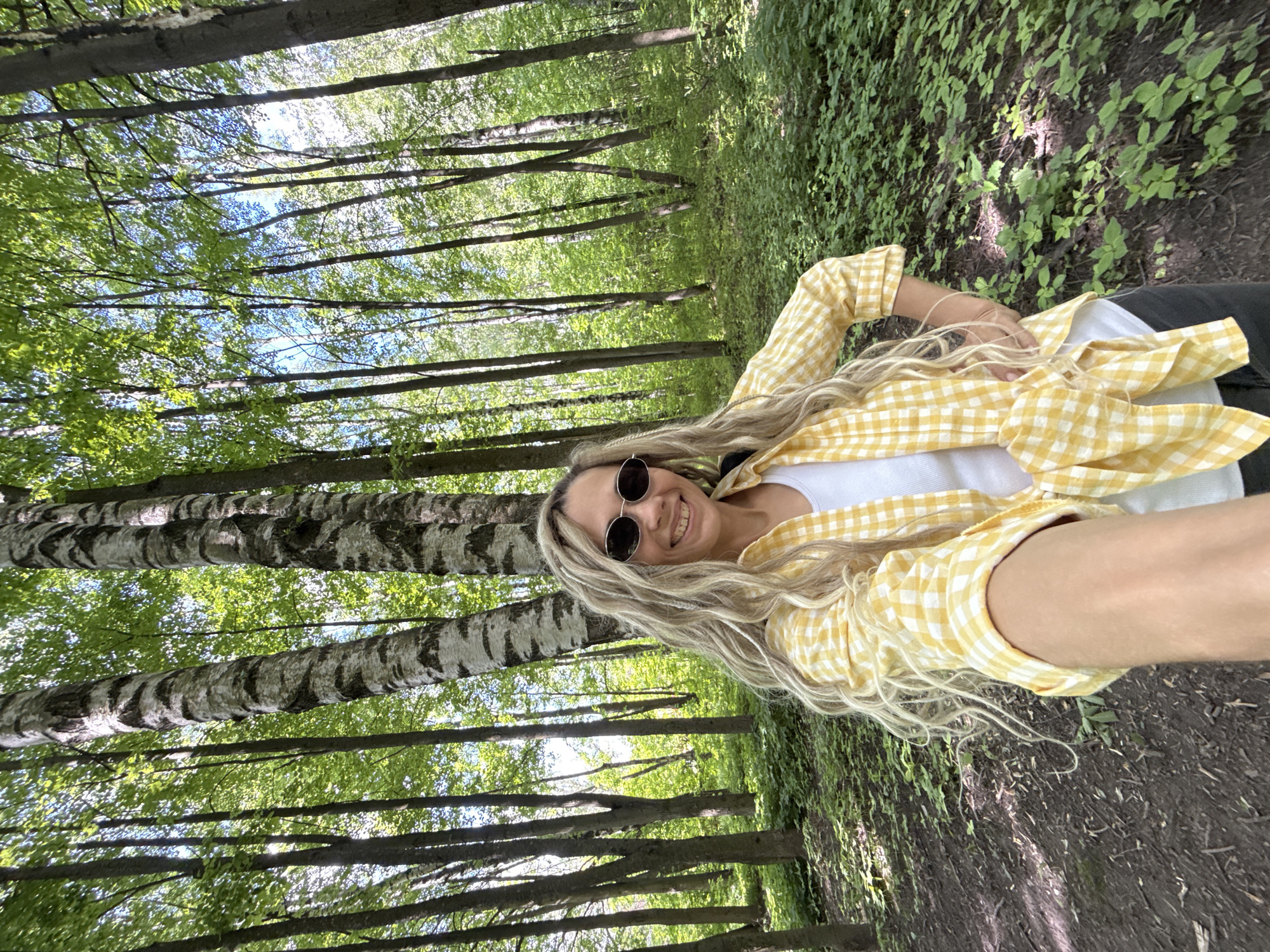 A medium-shot portrait captures a smiling woman taking a selfie in a sun-dappled forest, likely in Rusanovo, Russia. The image is oriented vertically, showing the woman's upper body and the surrounding environment.
The main subject is a woman with long, wavy blonde hair that cascades over her shoulders. She is wearing dark, round sunglasses, and a cheerful smile illuminates her face. Her attire consists of a yellow and white gingham (checkered) button-down shirt, worn open over a plain white top. One hand is placed casually on her hip.
The background is dominated by tall, slender tree trunks, some exhibiting distinctive white and dark striped bark, indicative of birch trees common in Russian forests. Lush green foliage fills the canopy above, with bright patches of blue sky visible through the leaves, suggesting clear and sunny weather. The forest floor is covered with a mix of leafy ground cover and dark soil, with hints of a path or trail. The lighting indicates it is daytime, with sunlight filtering through the dense leaves, creating a bright and pleasant atmosphere. No visible text is present in the image. The overall mood is joyful and serene, reflecting the woman's positive expression and the natural beauty of the forest.