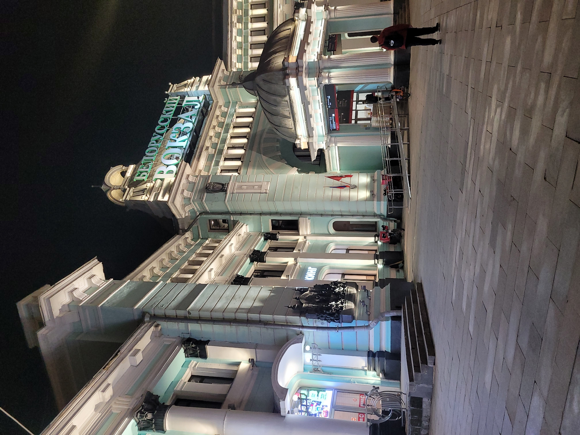 A nighttime photograph captures a corner view of the Belorussky Railway Terminal in Moscow, Russia. The architectural style includes light-colored facades, ornate details, and illuminated signage that reads "БЕЛОРУССКИЙ ВОКЗАЛ" in green neon letters. A pedestrian in a red jacket is walking along the paved pathway in front of the building. The sky is dark, and the building is well-lit, highlighting its architectural features.
