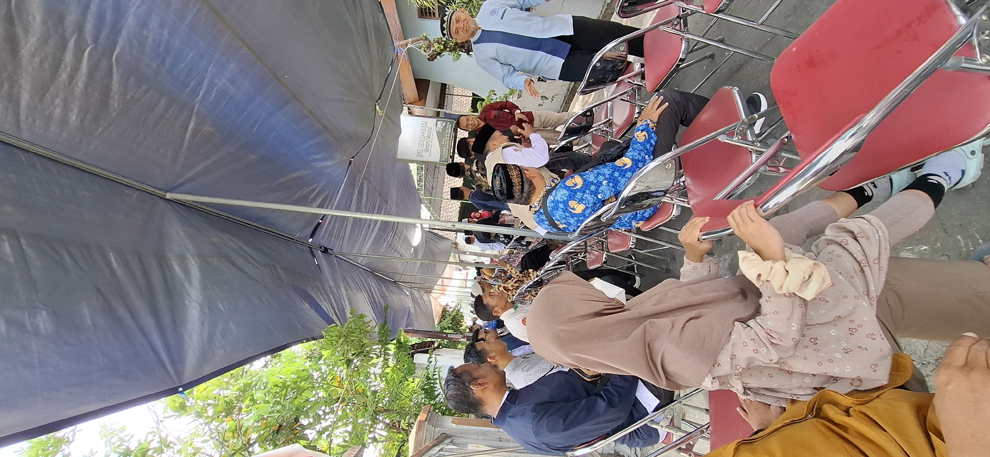 The image depicts an outdoor gathering under a dark blue tarp. Several people are seated on red chairs, some facing away from the camera and others partially visible. On the left, a man in a dark blue jacket and a woman wearing a light brown hijab and a patterned dress are seated. To their right, a man in a white shirt and another in a blue patterned shirt are also seated. In the background, more individuals are visible, some standing. To the right of the seated individuals, a man in a light blue and white collared shirt and black pants stands and gestures with his hands. A sign visible in the background reads "PT. ZAM-ZAM TOUR & TRAVEL". The environment appears to be a street or courtyard with a wall and some greenery visible on the left. The overall impression is a communal event, possibly a religious or cultural gathering, taking place in Gentan, Indonesia.