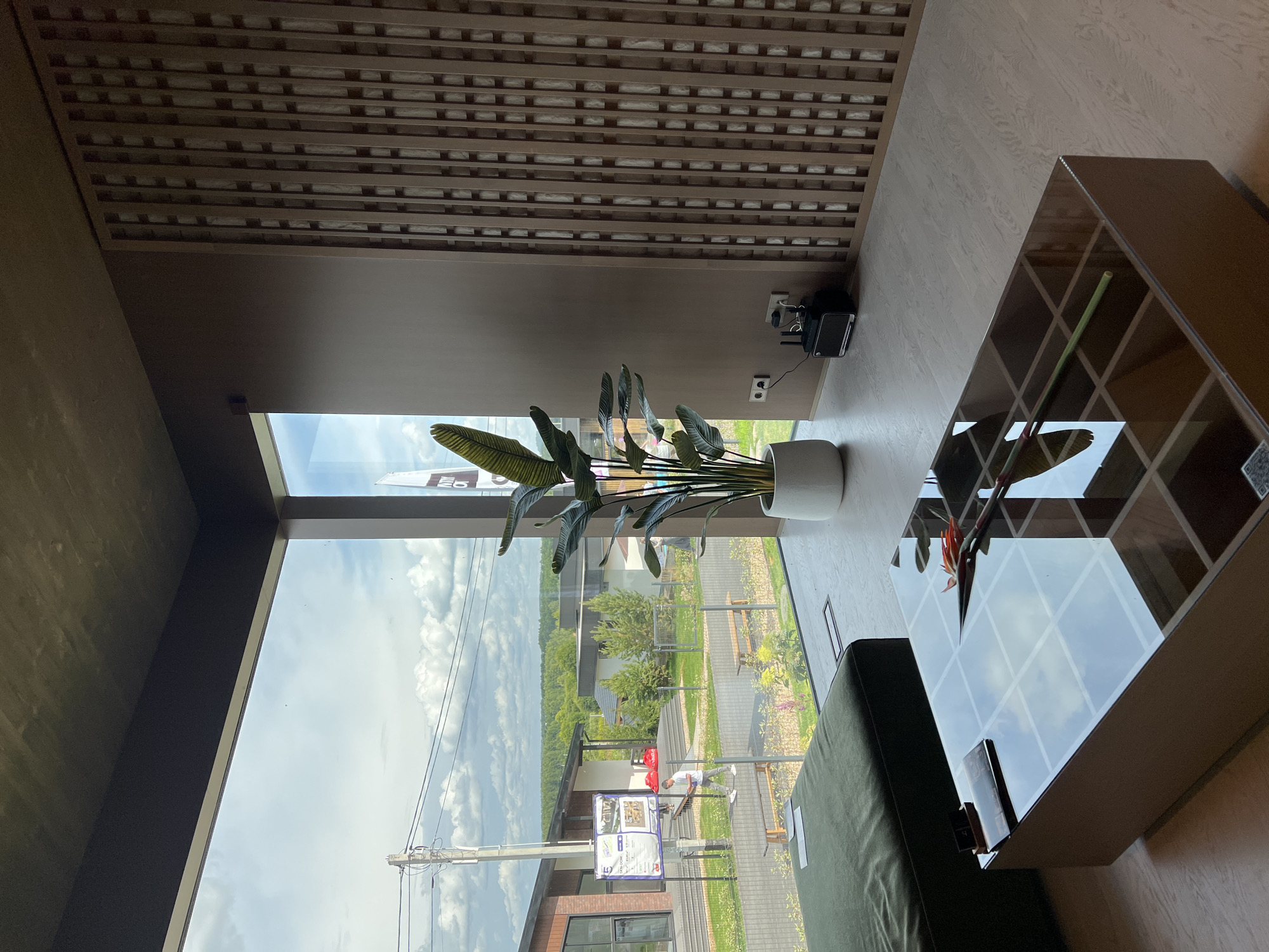 A high-angle interior shot captures a contemporary living space with a large window offering a view of a suburban landscape in Yakunino, Russia. The room features a dark green ottoman on the left, a glass-topped coffee table in the foreground with a flower and a book, and a large potted plant in a white pot on the right. A wooden slatted wall design is visible on the far right. Through the window, houses, trees, a cloudy sky, and a utility pole are visible. A person walks by the houses outside, and a signpost stands near the entrance of one of the houses. A small black speaker is placed on the floor near the plant.