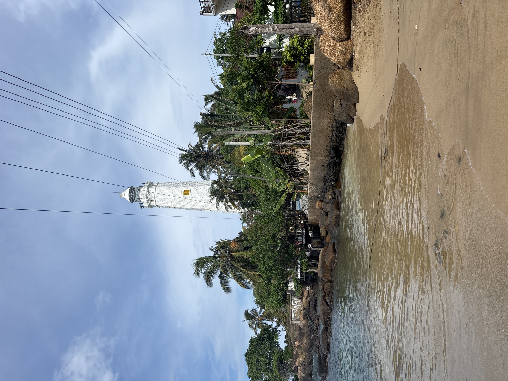 The image shows a coastal scene in Dondra, Sri Lanka. A white lighthouse stands tall amidst palm trees and other vegetation. Utility wires are visible against a partly cloudy sky. The foreground features a beach with sand and water, with a rocky embankment separating the land from the sea. The image is oriented sideways.