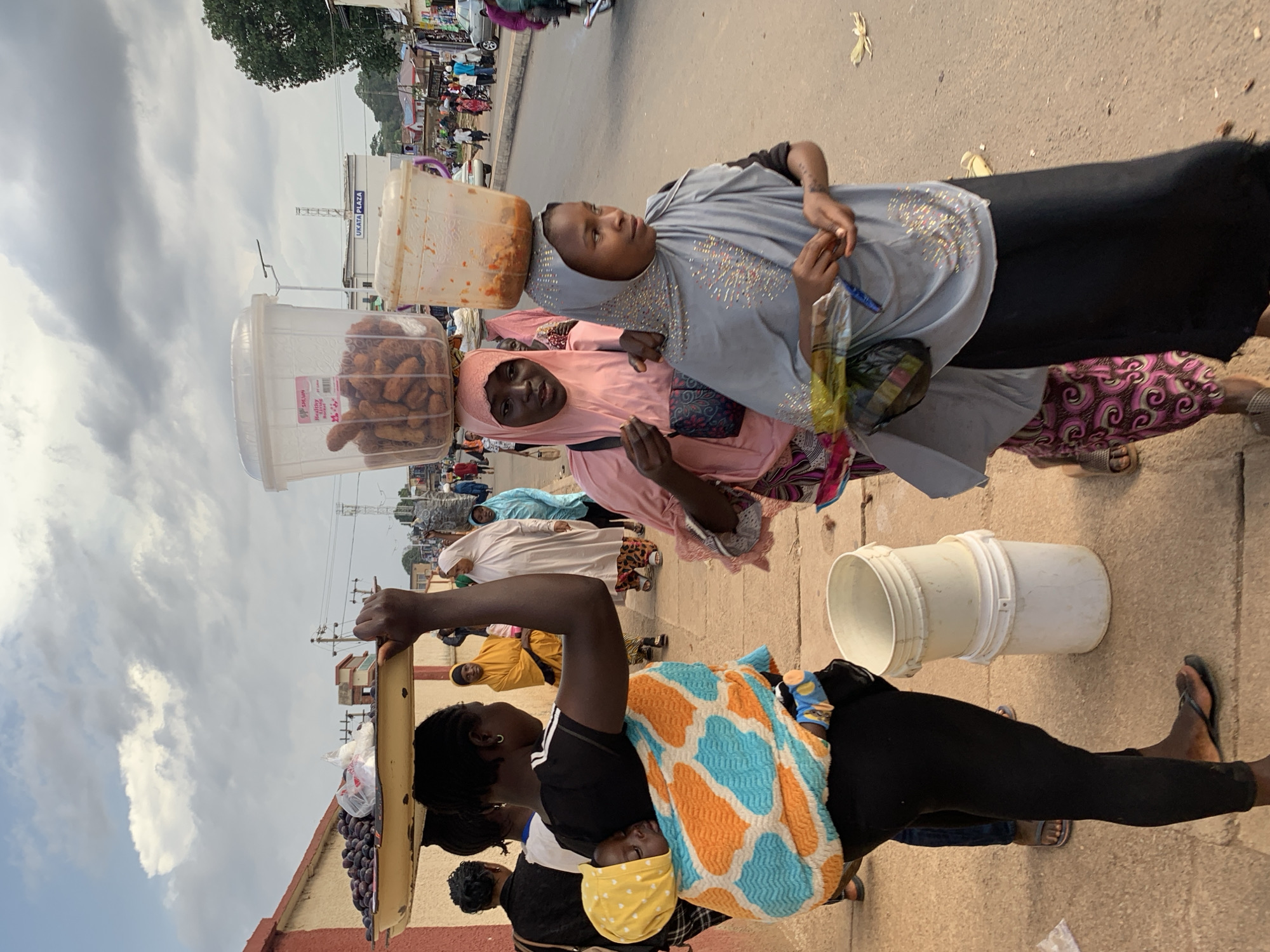 The image depicts a busy street scene, likely a marketplace, in Jos, Nigeria. The primary subjects are several women, some of whom are carrying large buckets balanced on their heads. One woman in the foreground is wearing a black top with white stripes on the sleeves and a colorful blanket wrapped around her, with a baby also wrapped in the blanket. Another woman next to her is wearing a pink hijab and a pink top, and she is also carrying a bucket on her head, filled with what appear to be fried snacks. A third woman, further back, is wearing a light blue hijab and a grey top adorned with rhinestones, also carrying a bucket on her head.
The objects visible include buckets, a tray filled with dark purple grapes, and plastic bags containing food items. The setting is an outdoor street with a paved surface. In the background, there are buildings and other people walking around, suggesting a lively market environment. There are also power lines and a tall structure with the text "UKATA PLAZA" visible on a building.
The time of day appears to be daytime, with natural light illuminating the scene. The sky is partly cloudy. The overall impression is one of a bustling, everyday market activity in Nigeria, with women engaged in commerce and daily life. The activity is focused on the sale and transport of goods, specifically food items. The expressions on the faces of the women are neutral, but the overall atmosphere suggests a sense of purpose and routine.