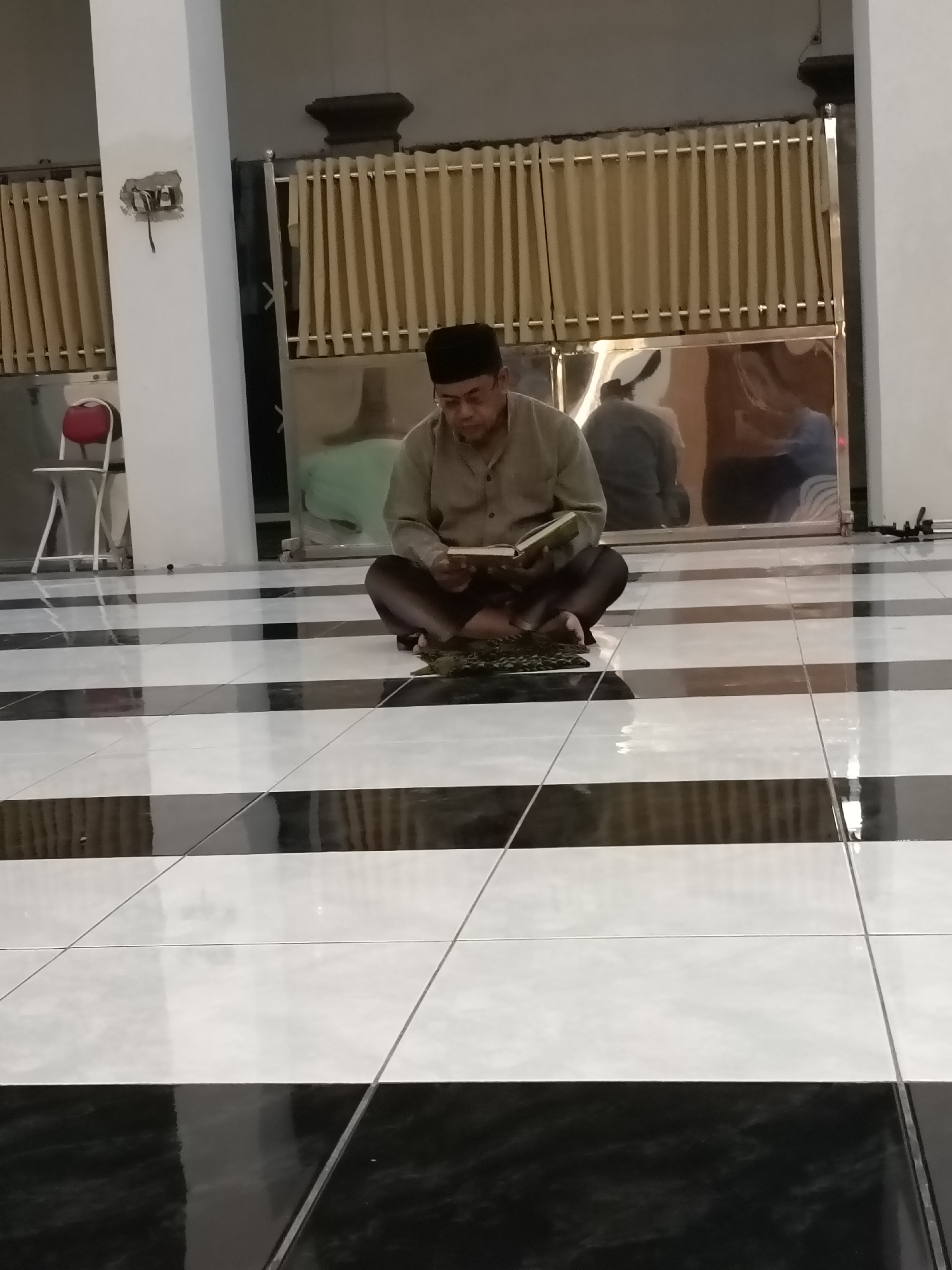 A man wearing a black cap and a light brown long-sleeved shirt sits cross-legged on a black and white tiled floor in what appears to be a prayer room or mosque. He is holding an open book and appears to be reading. A dark green prayer mat is placed in front of him. The room has beige curtains in the background, and a reflective surface that shows the blurred figures of other people. A white pillar with exposed electrical wiring is visible to the left, along with a red-seated chair. The overall scene suggests a quiet moment of devotion. The location is the city of Kediri, Indonesia.